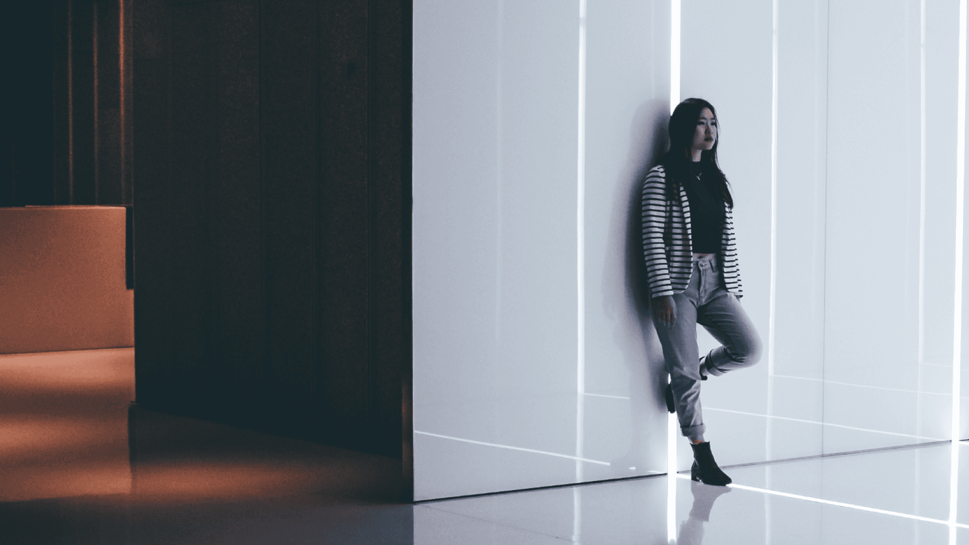 Woman in striped jacket leans against a white, illuminated wall in a modern, dimly lit space, with reflective flooring.