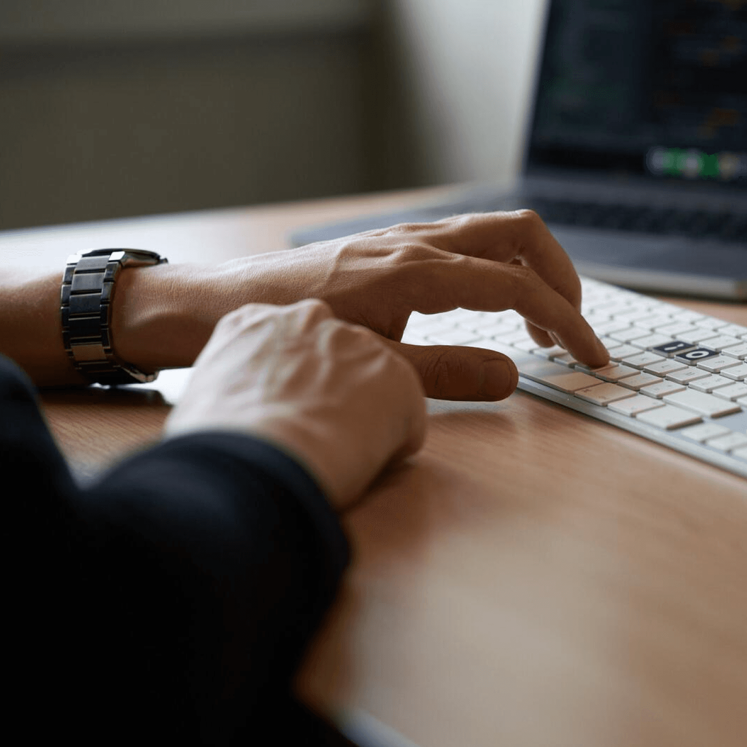 Hands typing on a wireless keyboard with a laptop in the background on a wooden desk.