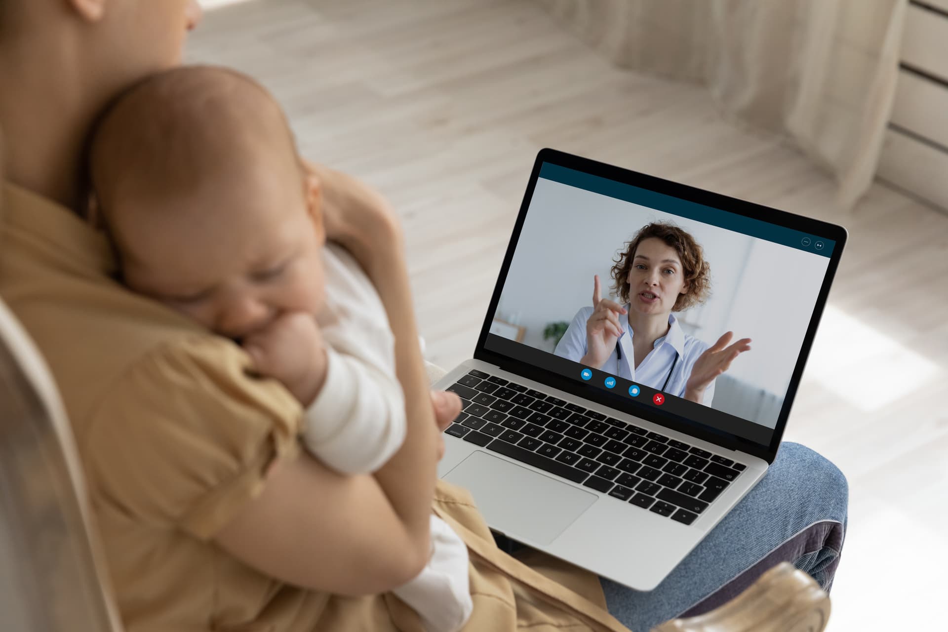 A woman holds a baby while having a video call on a laptop with a doctor, who is gesturing and speaking.