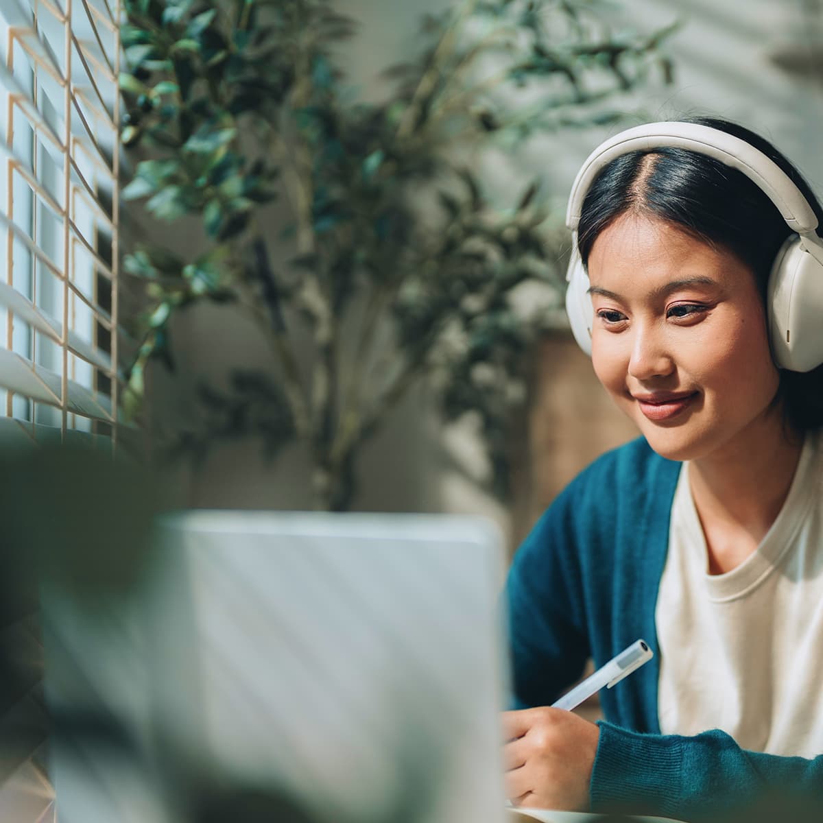 A person wearing headphones, smiling and writing, sits in front of a laptop with plants and sunlight streaming through window blinds in the background.