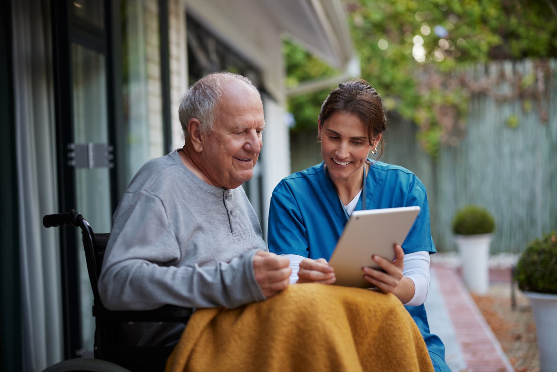 Elderly man in a wheelchair and a nurse with an iPad, smiling together in a garden setting.
