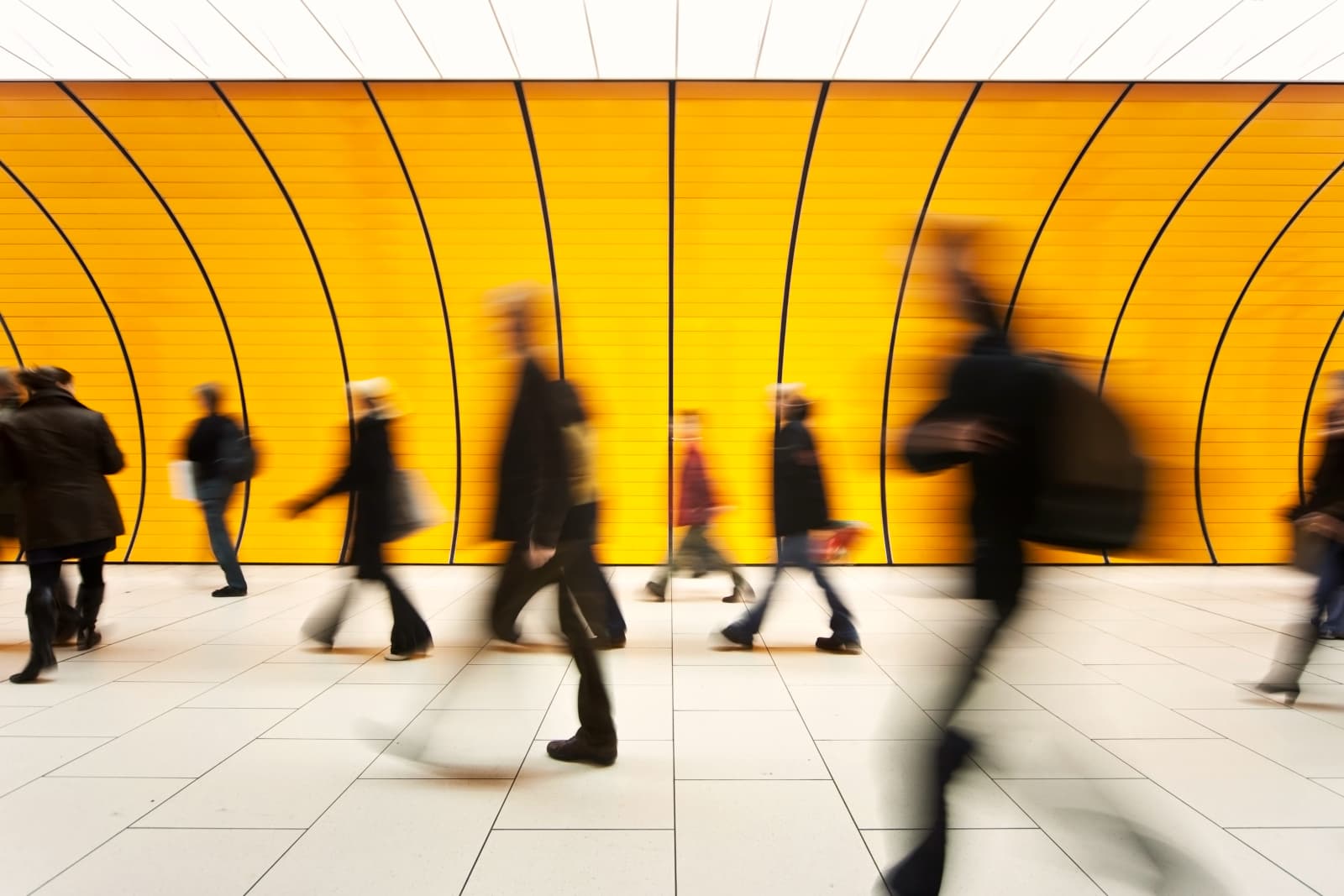 Blurry silhouettes of people walking past a bright yellow, curved wall in a modern, spacious corridor.