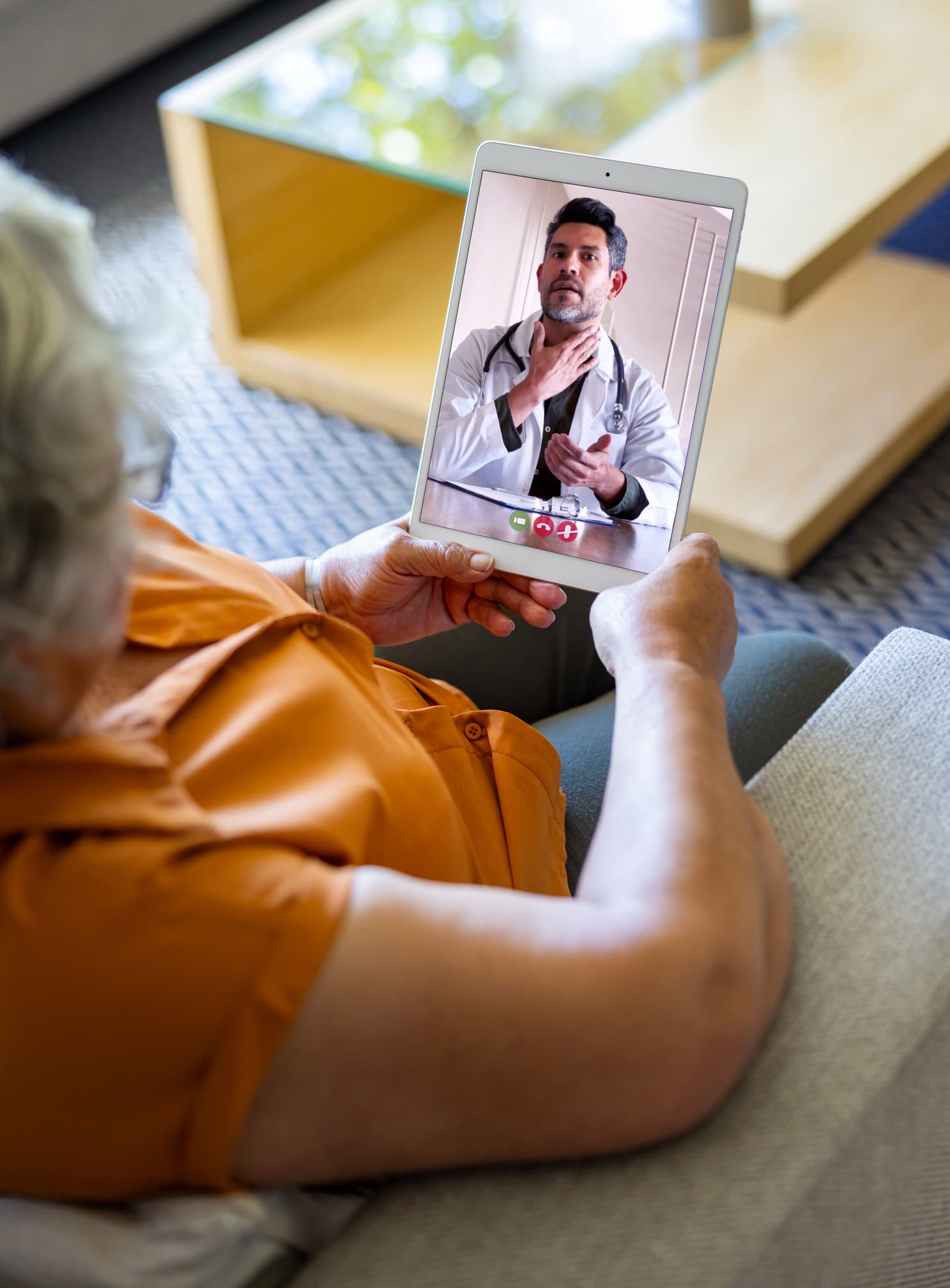 An elderly person video calls a doctor on a tablet, with the doctor gesturing to his throat, possibly discussing symptoms or health advice.
