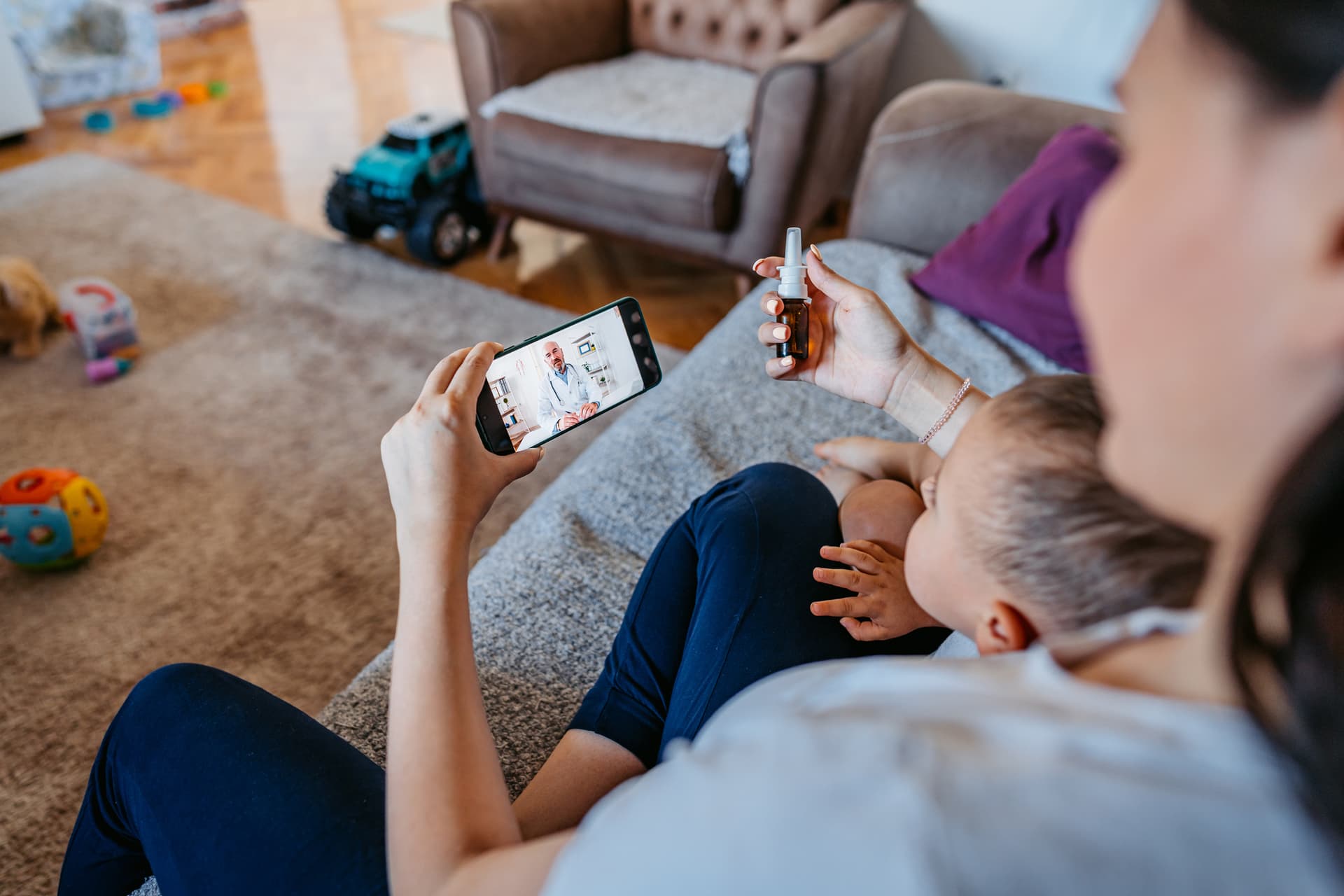 A woman video chats on a smartphone while holding a baby and a nasal spray, seated on a couch in a cozy living room with toys on the floor.