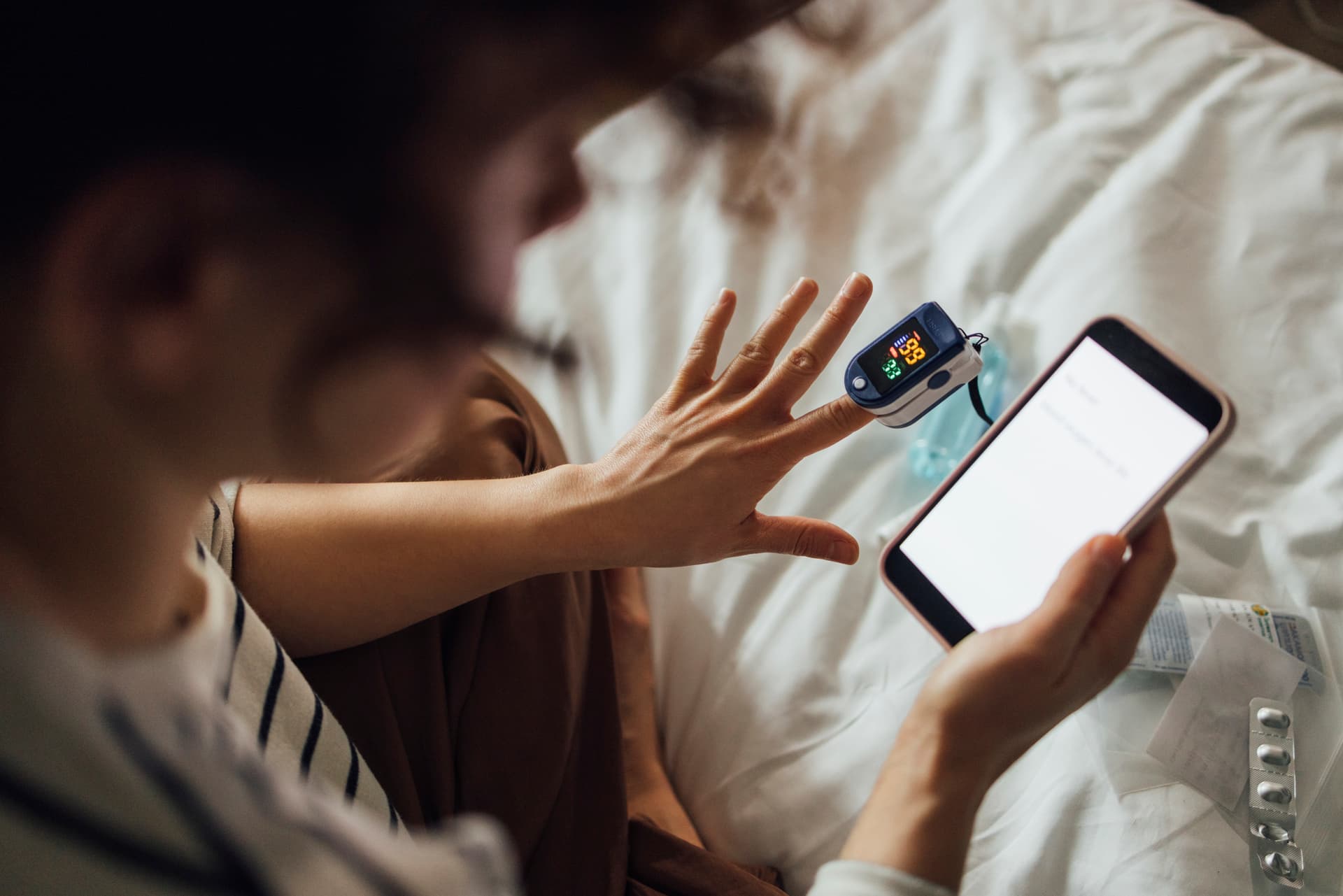 Person checking oxygen saturation with a finger pulse oximeter while holding a smartphone, sitting on a bed.