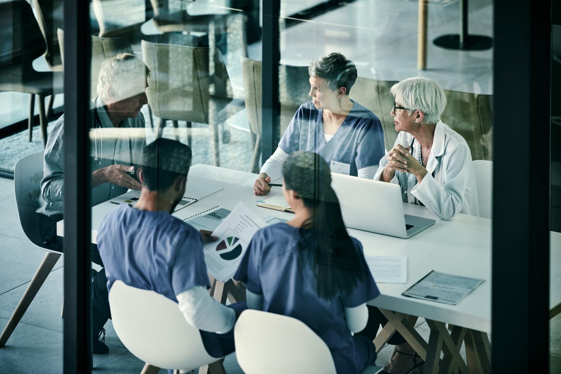 A diverse group of medical professionals in a glass-walled conference room discussing charts and data around a table with laptops and documents.