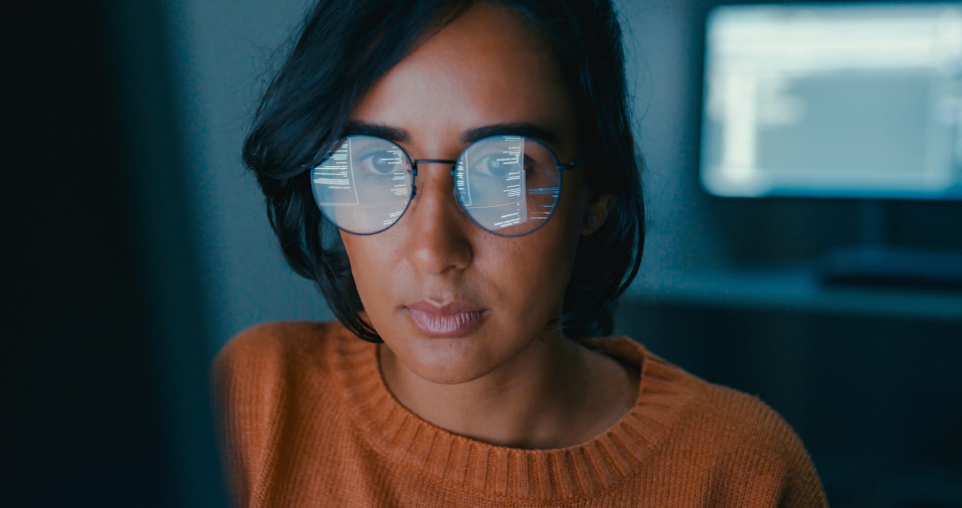 Person wearing glasses, focused on a computer screen with code reflecting in the lenses, in a dimly lit room.