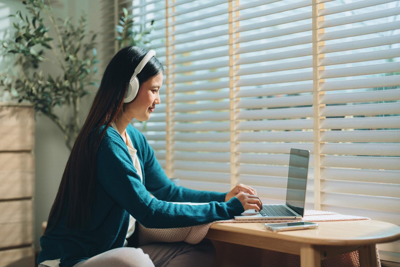 A woman wearing headphones works on a laptop at a wooden table, with sunlight streaming through window blinds.