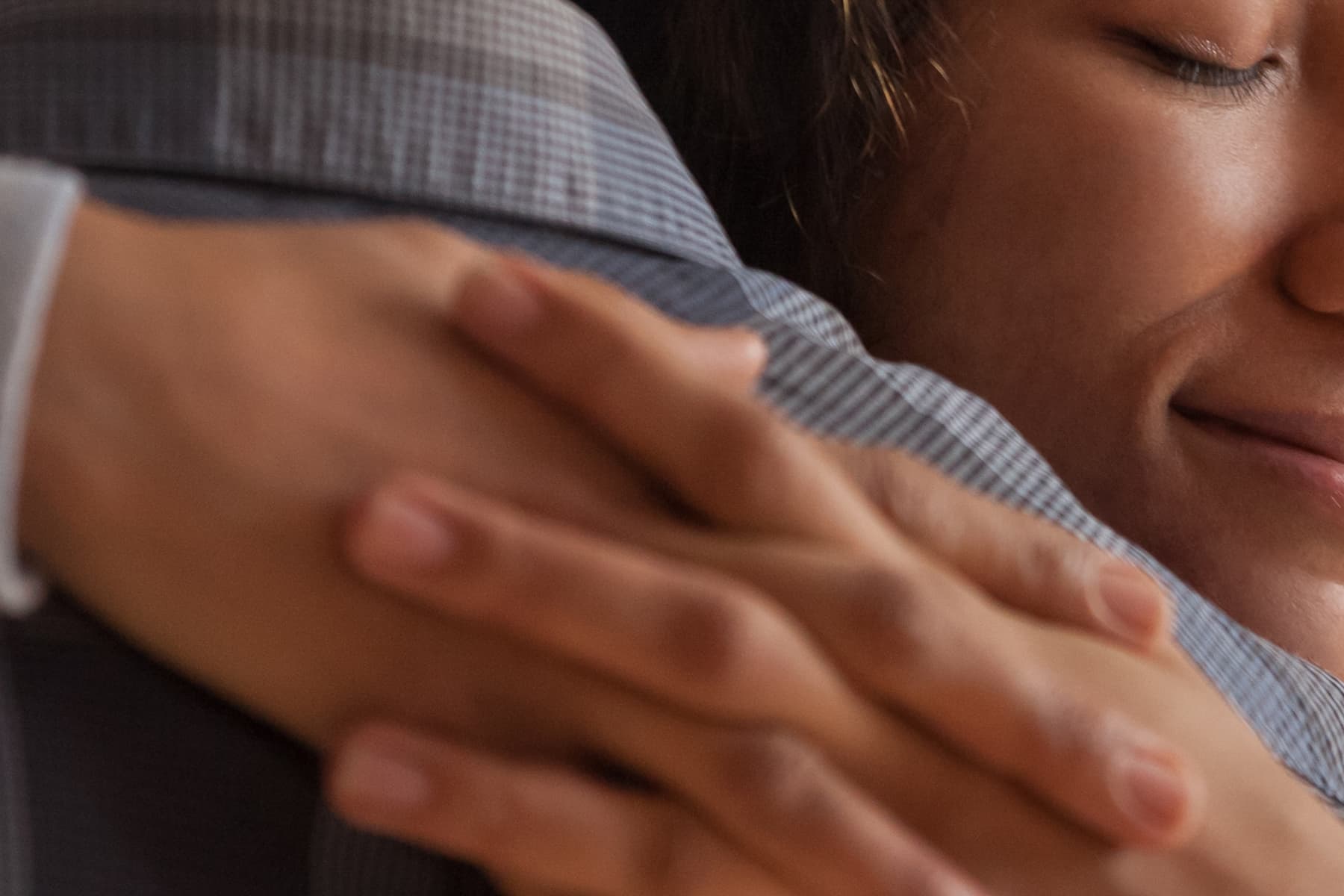 Close-up of two people hugging, showing intertwined hands and a smiling face. One person wears a gray suit.