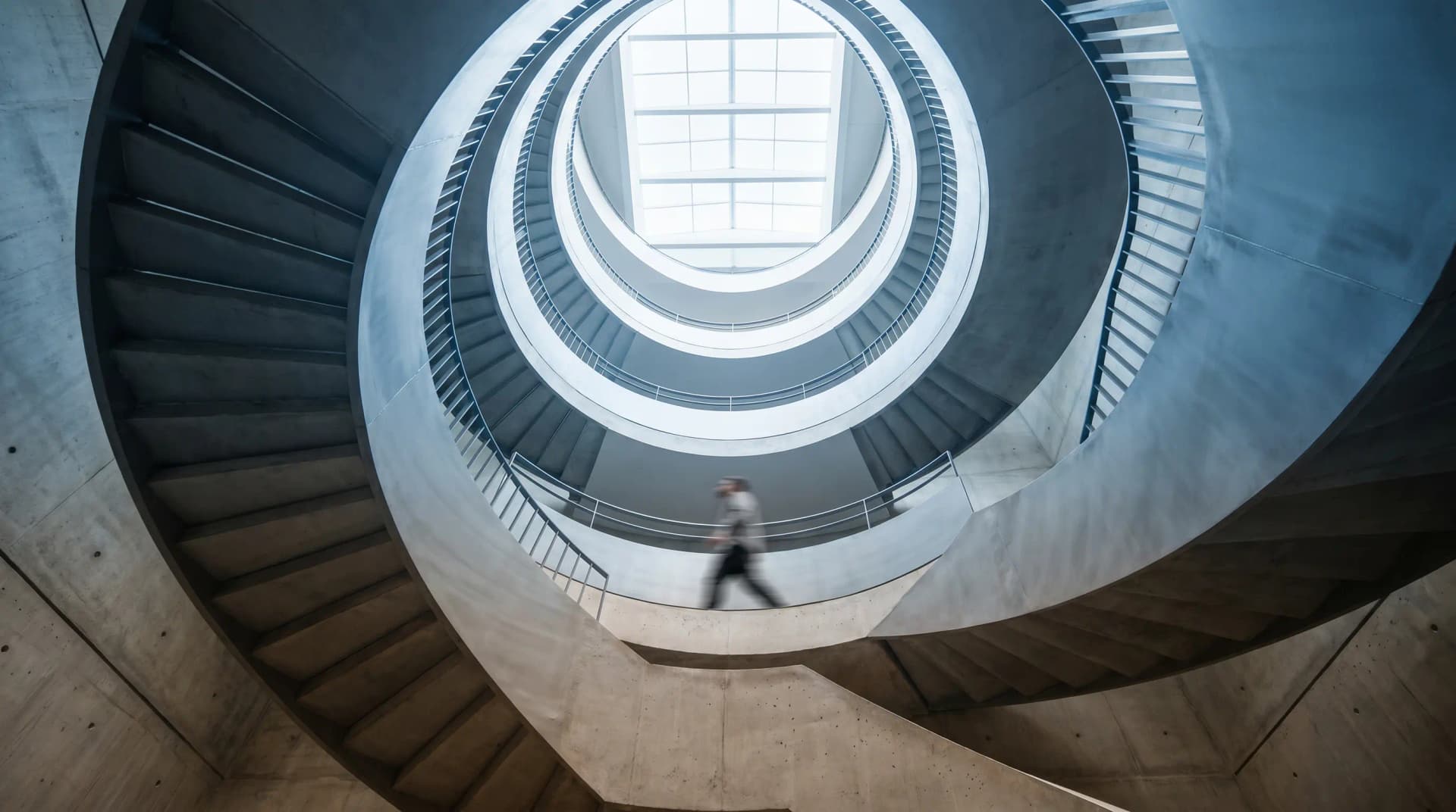 Spiral staircase with a blurred person walking, viewed from below, featuring concentric circular patterns and a skylight above.