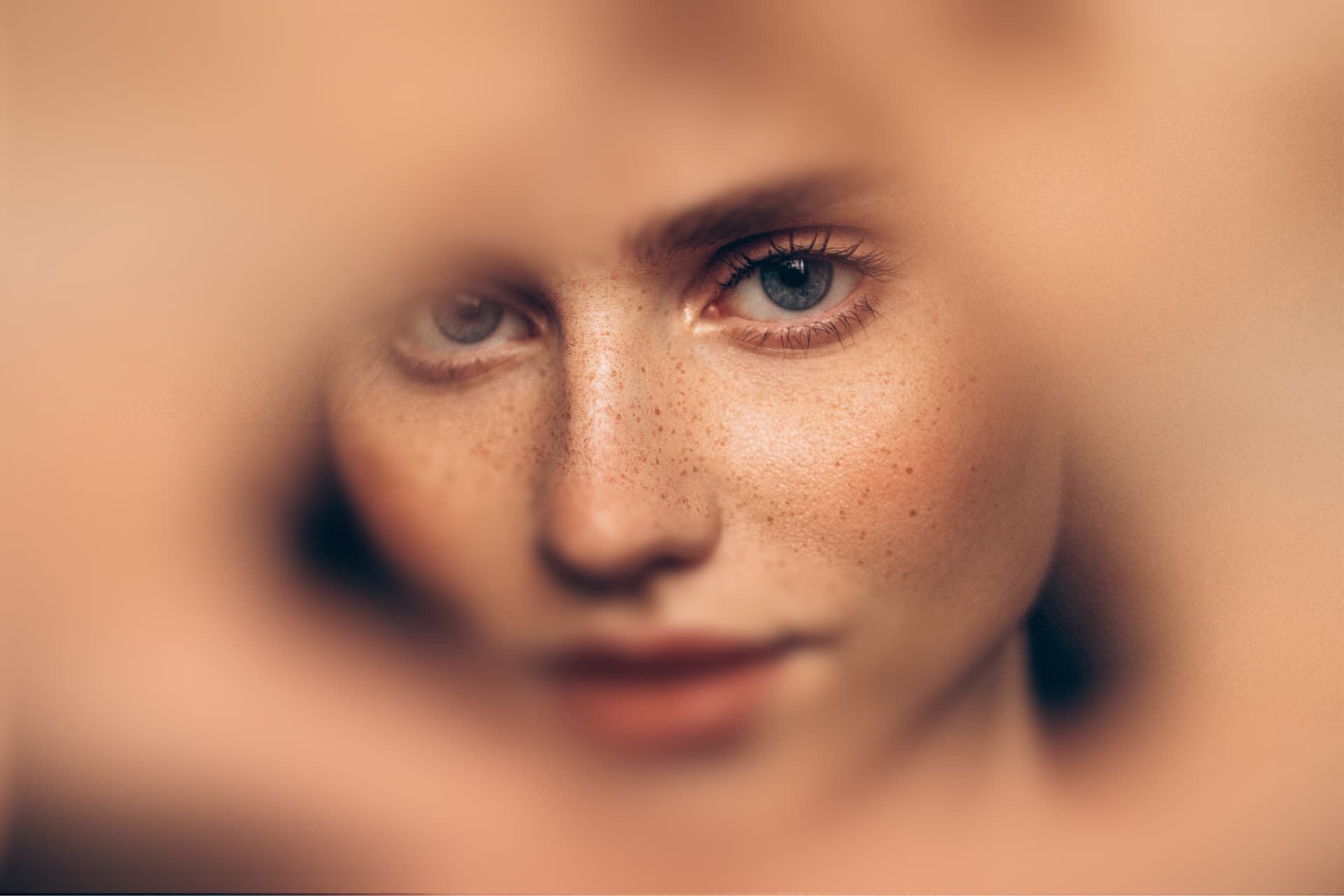 Close-up of a person with blue eyes and freckles, framed by blurred hands. The focus is on the face, creating an intimate and artistic effect.
