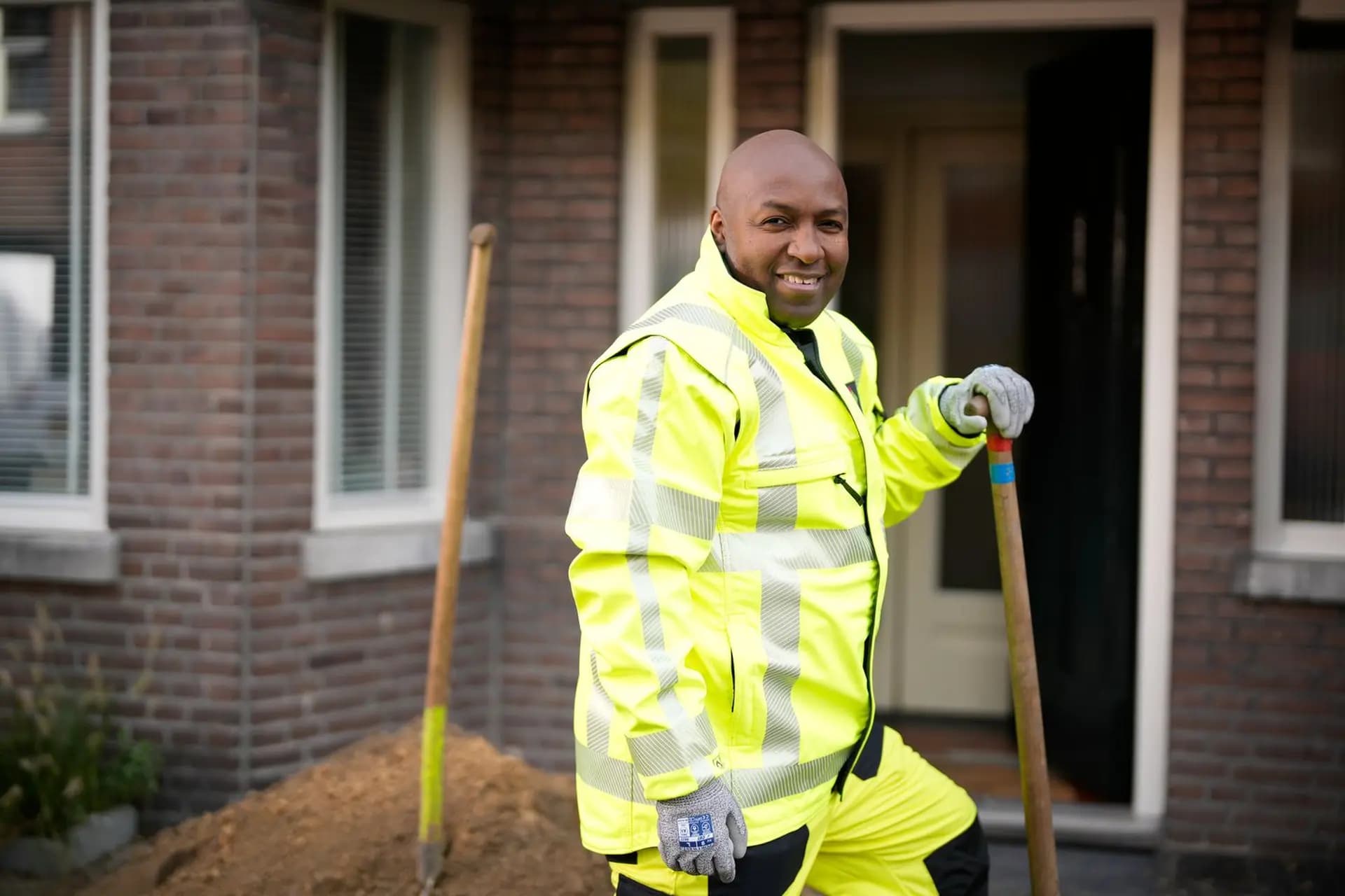 Smiling man in a bright yellow reflective jacket holding a shovel, standing near a pile of soil in front of a brick building.