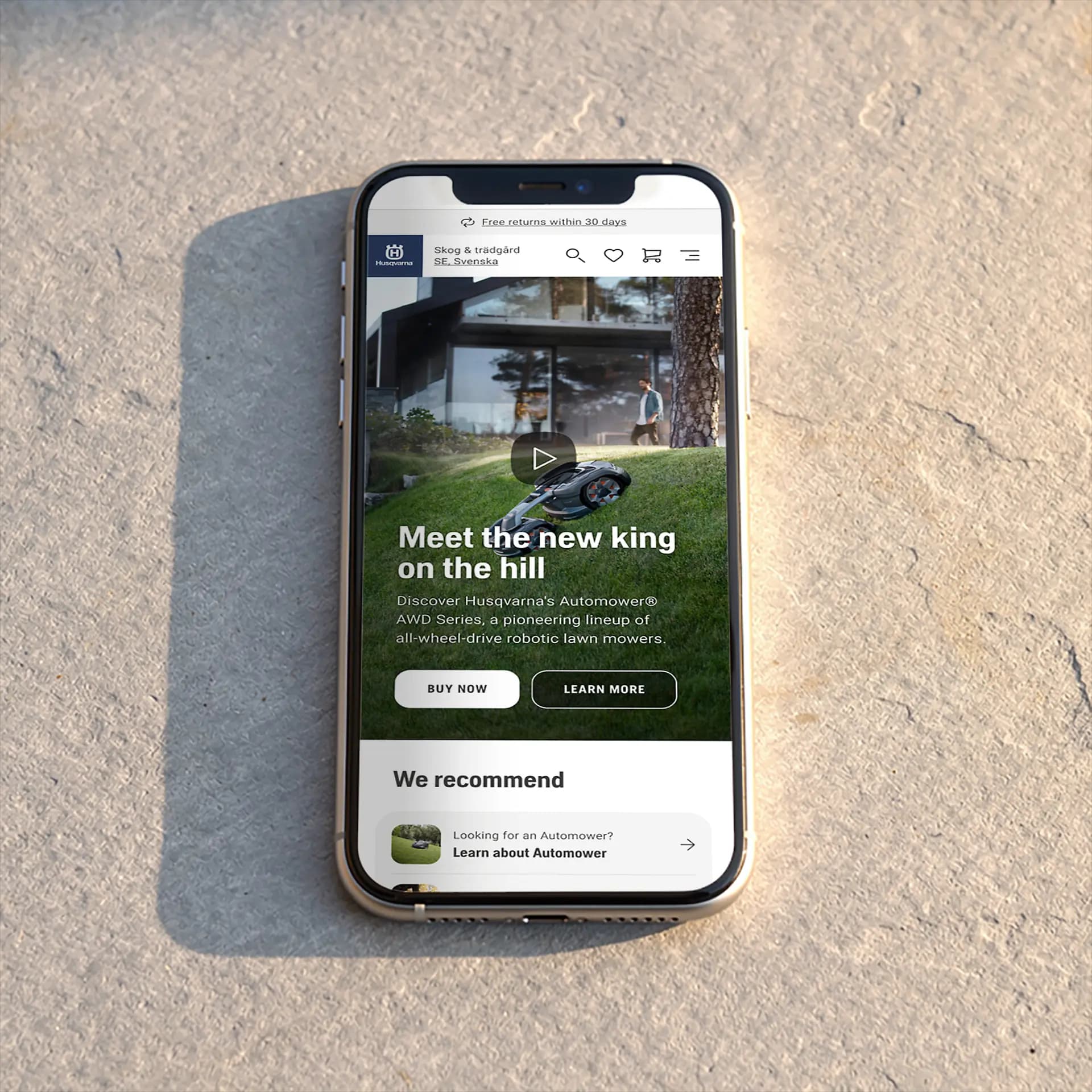 Smartphone on a stone surface displaying a webpage for Husqvarna's Automower, promoting robotic lawn mowers with "Buy Now" and "Learn More" buttons.