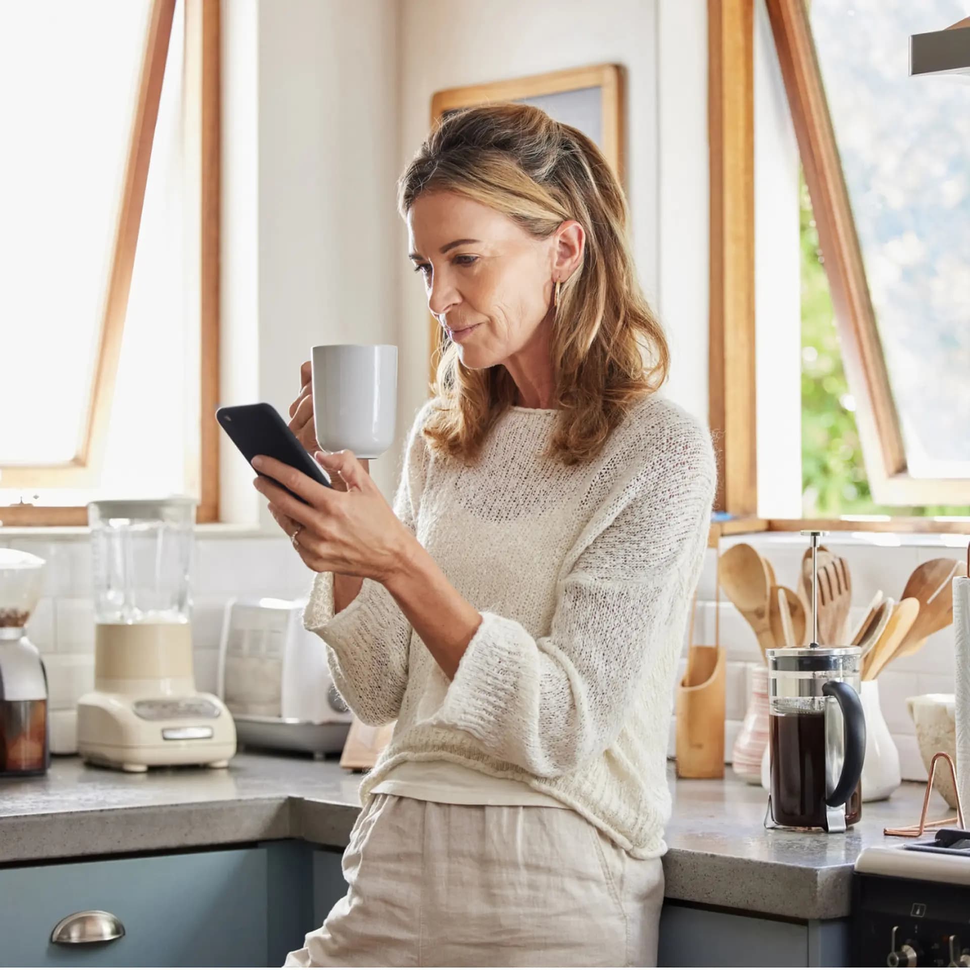 A woman in a kitchen sips from a mug while looking at her smartphone, surrounded by appliances and utensils, with sunlight streaming through the window.