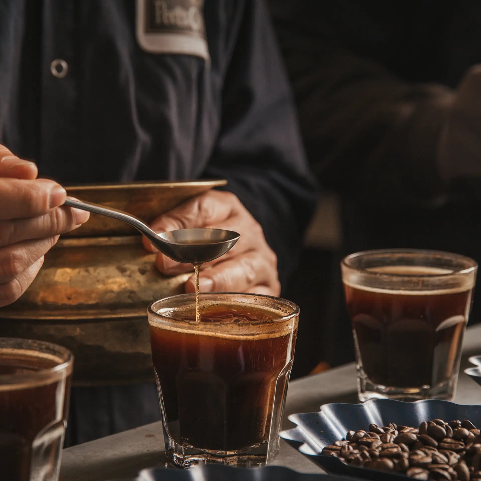 A person carefully pours liquid from a spoon into a glass of coffee, surrounded by other glasses and coffee beans on a table.