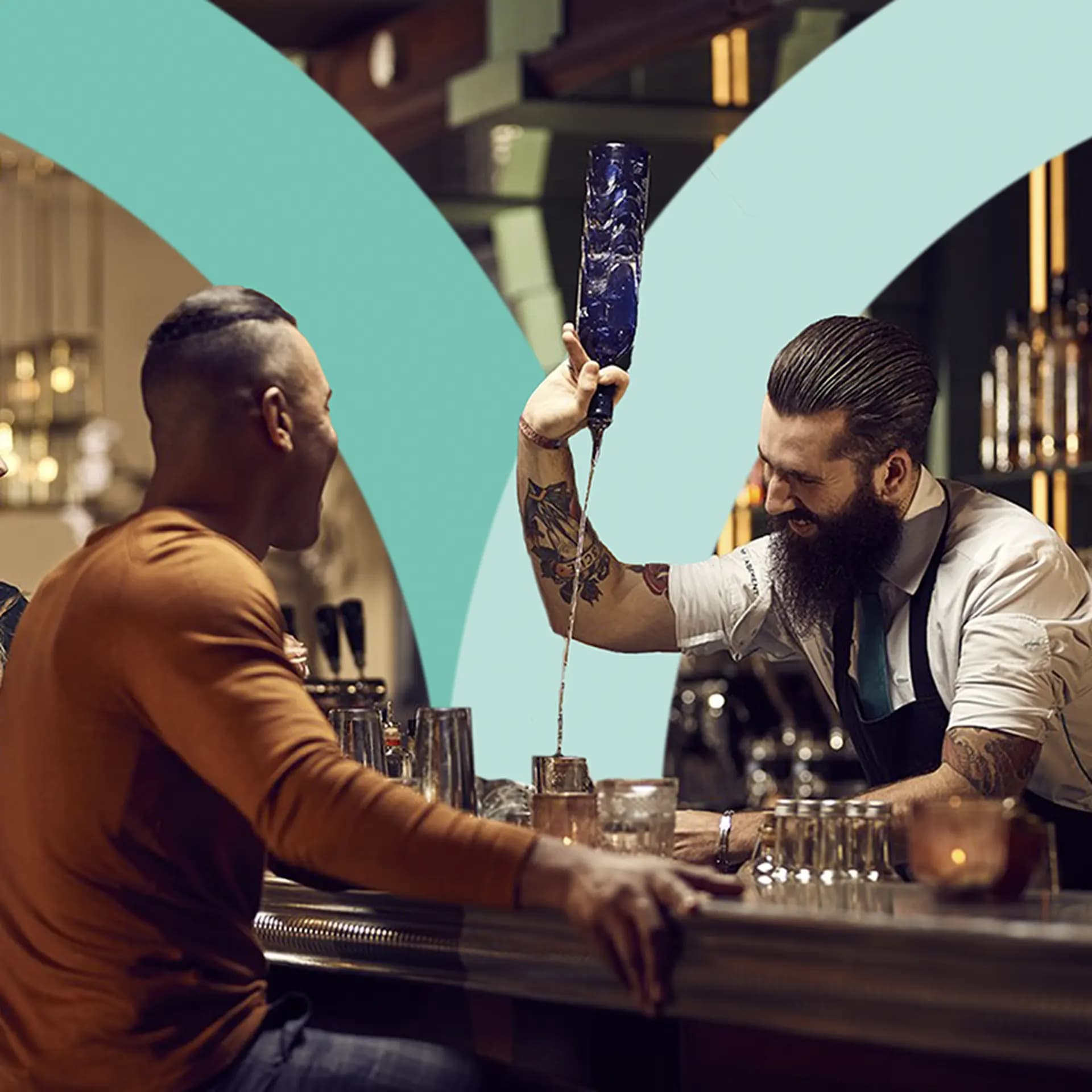 A bartender with a beard and tattoos pours a drink from a blue bottle, engaging with a smiling customer seated at the bar.