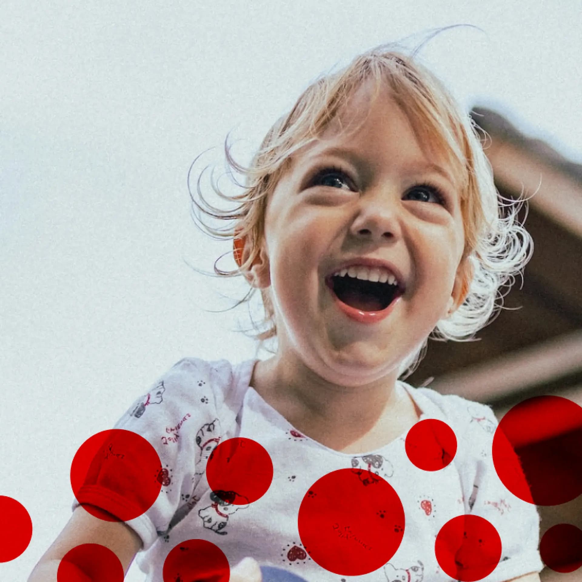 A laughing child with blond hair looks up, wearing a white patterned shirt. Red circles artistically overlay the image.