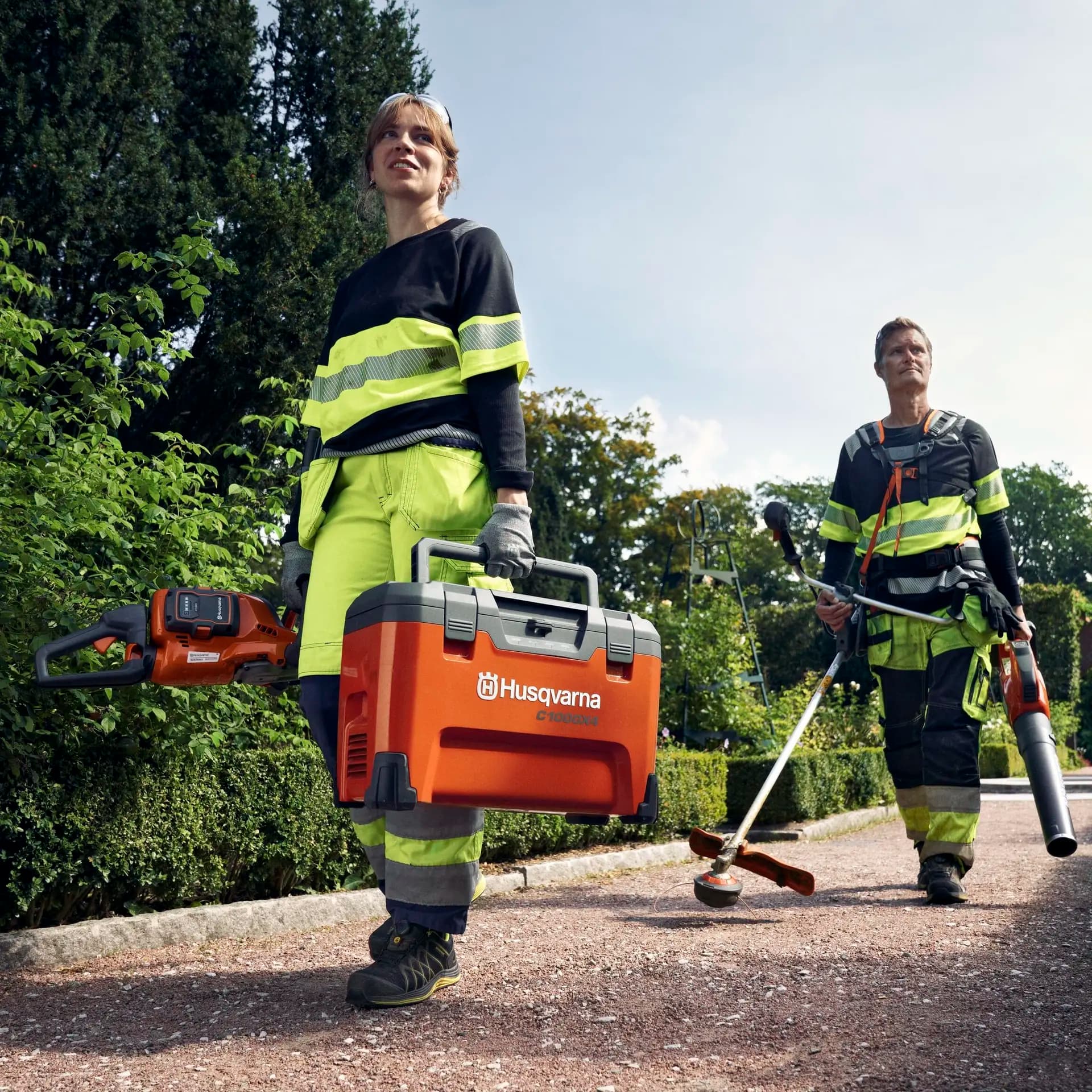 Two landscapers in fluorescent gear carry Husqvarna tools along a garden path, surrounded by lush greenery and a clear sky.