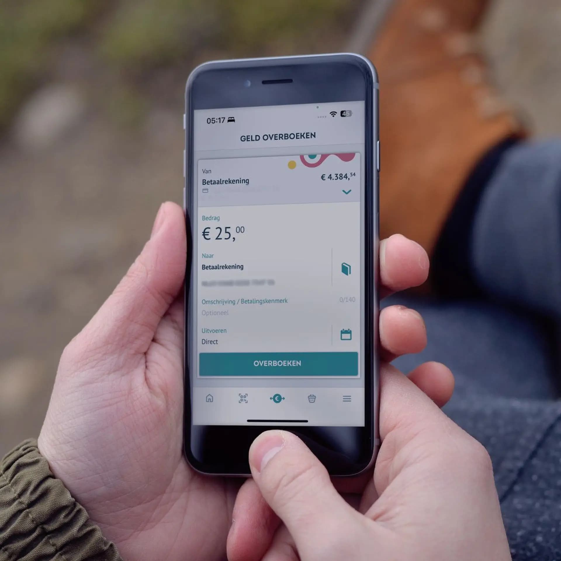 A person holding a smartphone displaying a banking app interface, showing a money transfer of €25 with the account balance of €4,384.54.