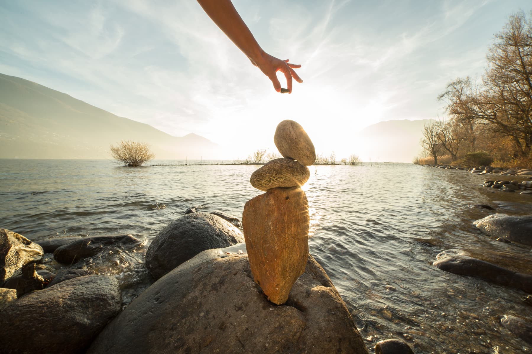 A hand balances stones on a lakeshore rock at sunset, with mountains and trees in the background.