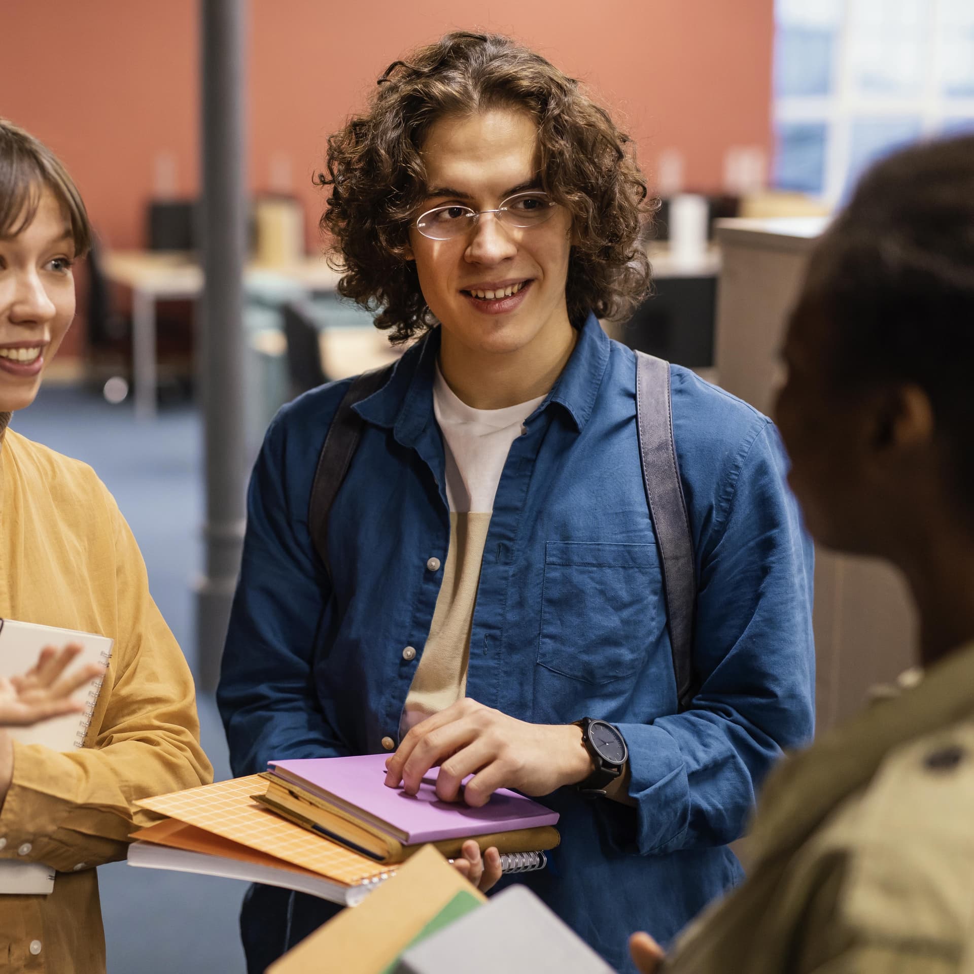 Three students chatting in a library, holding notebooks and smiling, with bookshelves and tables in the background.