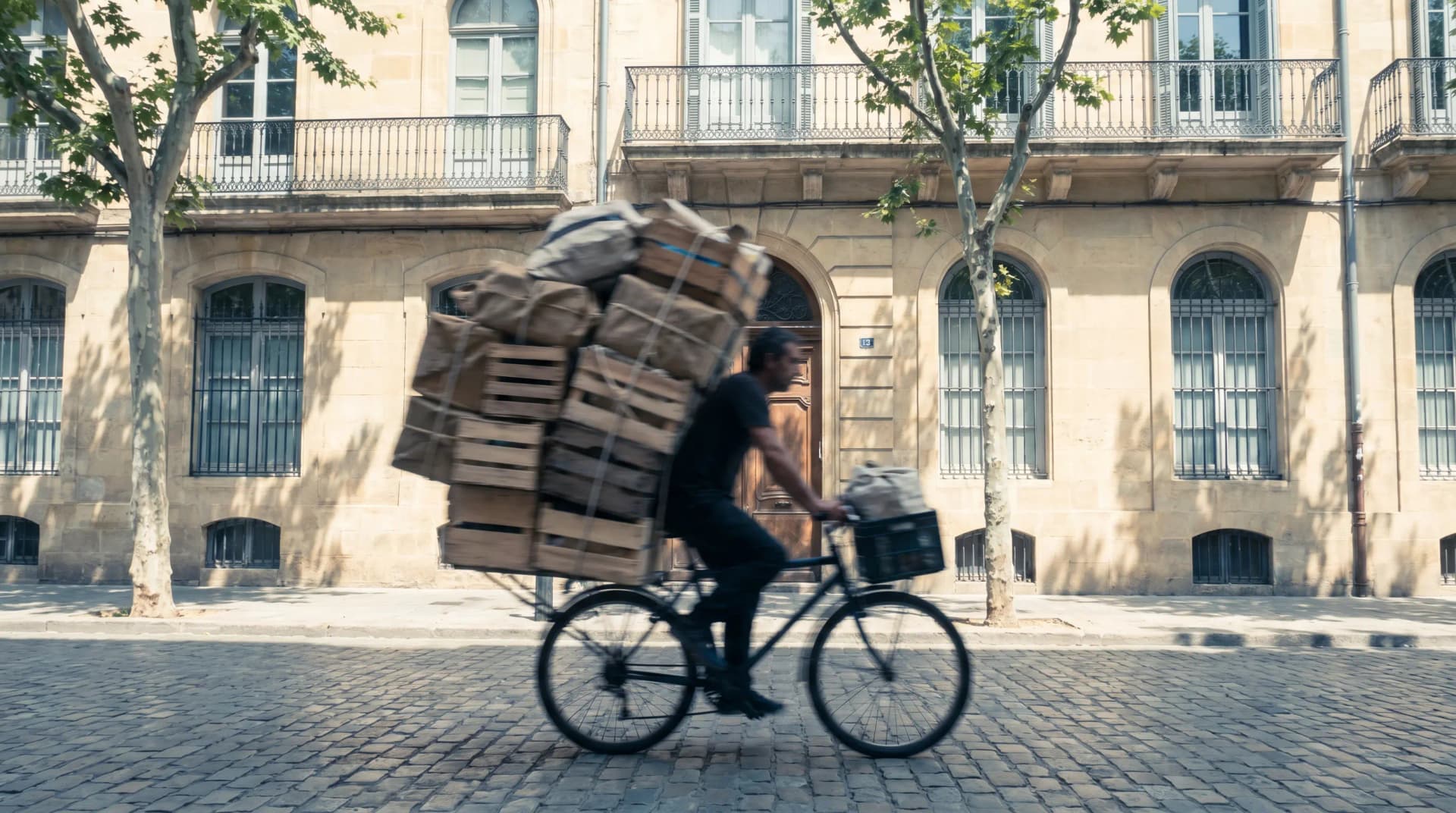 Person riding a bicycle loaded with boxes and crates, passing a historic building on a sunny cobblestone street.