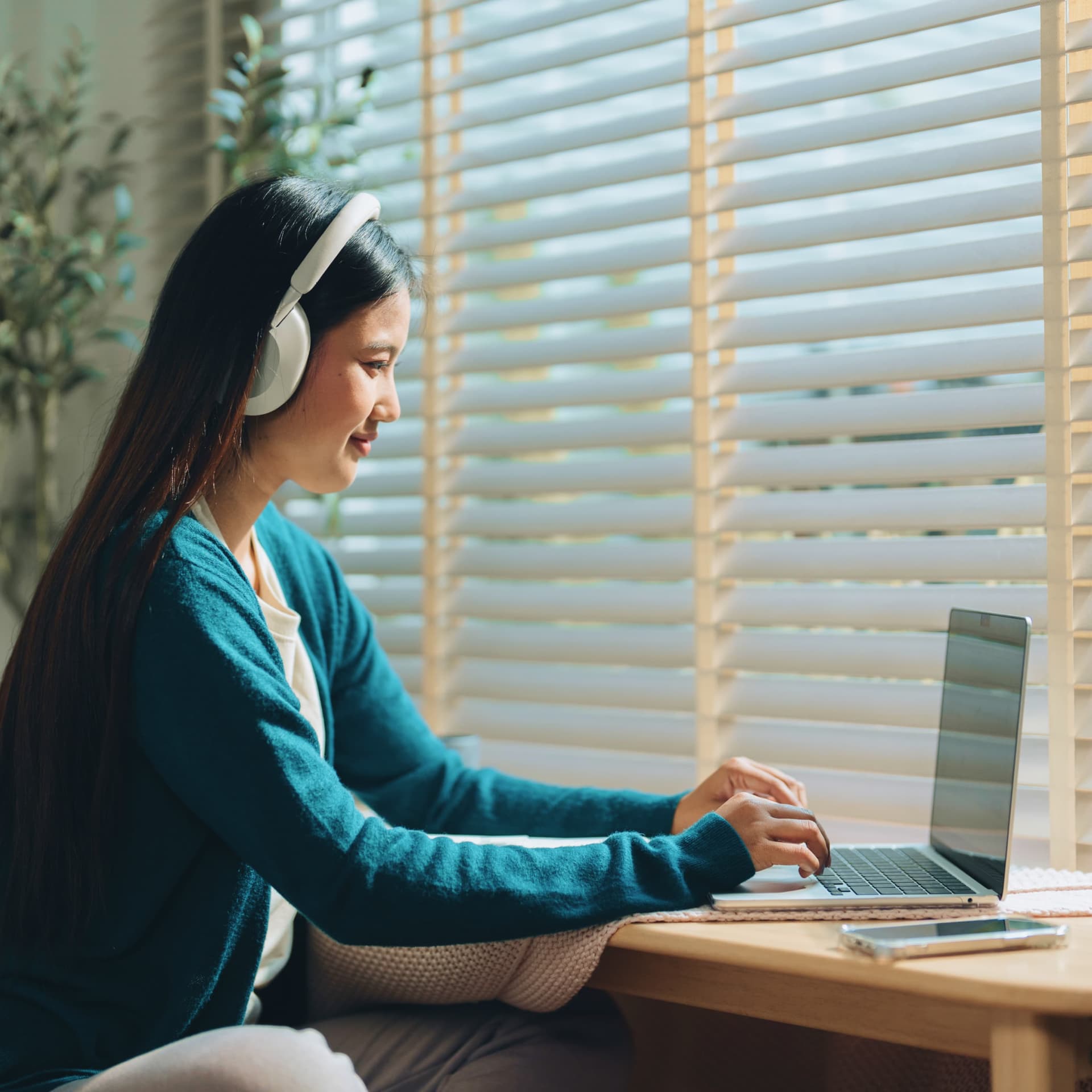 A woman wearing headphones sits by a window with blinds, working on a laptop at a wooden desk, looking focused and content.