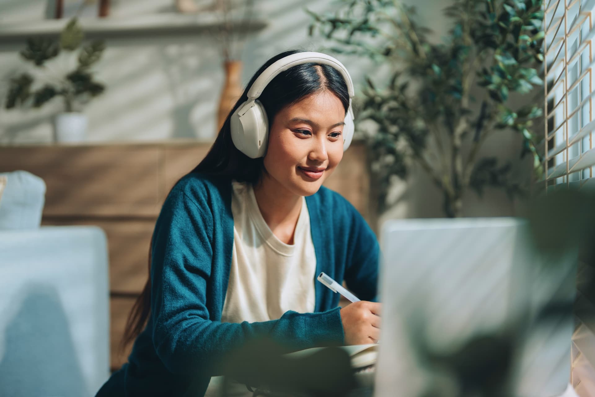 A woman in a blue cardigan, wearing headphones, writes notes while looking at a laptop in a cozy, plant-filled room.
