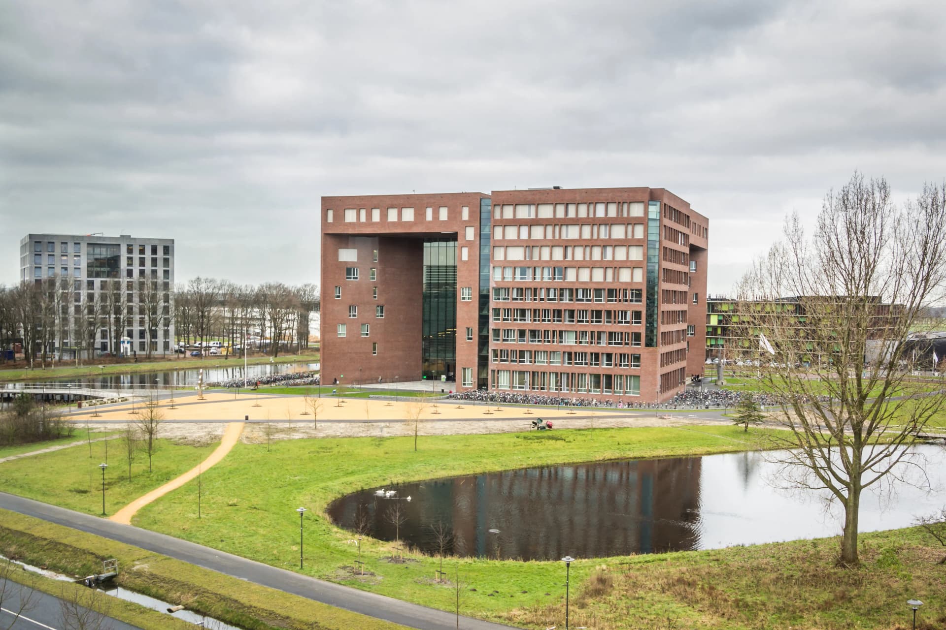 Modern brick building with large windows by a pond, surrounded by grassy landscape and pathways under a cloudy sky.