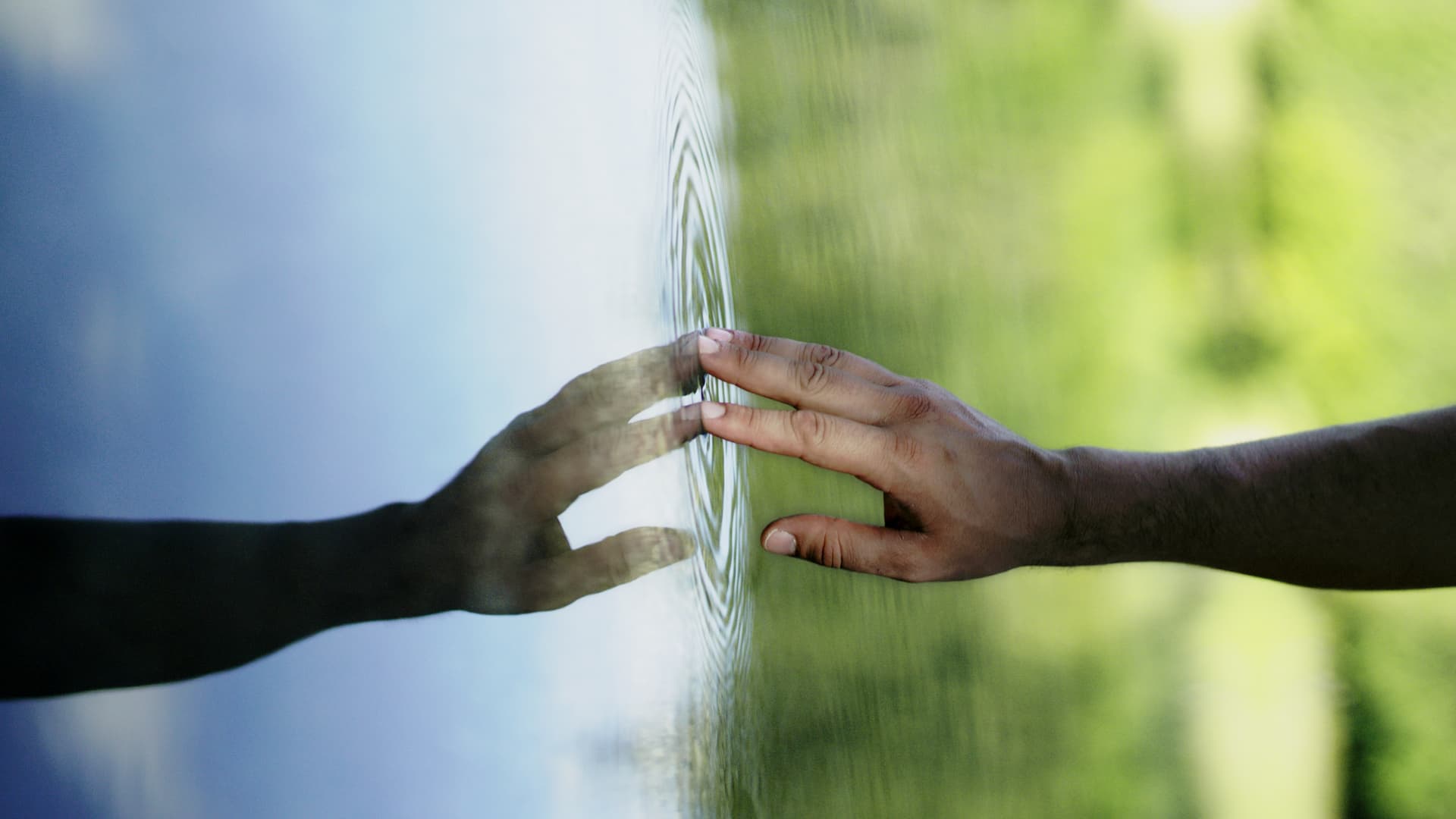 A hand touching the calm surface of water, creating ripples, with its reflection visible. The background is a blend of blue and green hues.