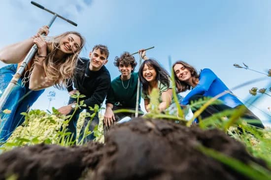 Five people smiling and leaning towards the camera while standing on grass, each holding a garden tool, with a bright blue sky above.