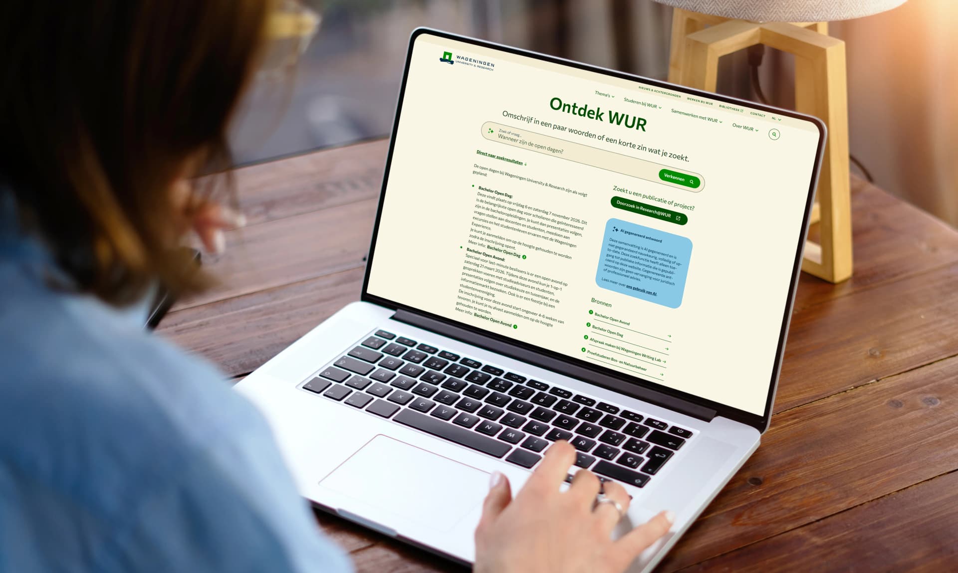 Person using a laptop displaying a webpage titled "Ontdek WUR" with text and search options, sitting at a wooden table with a lamp nearby.