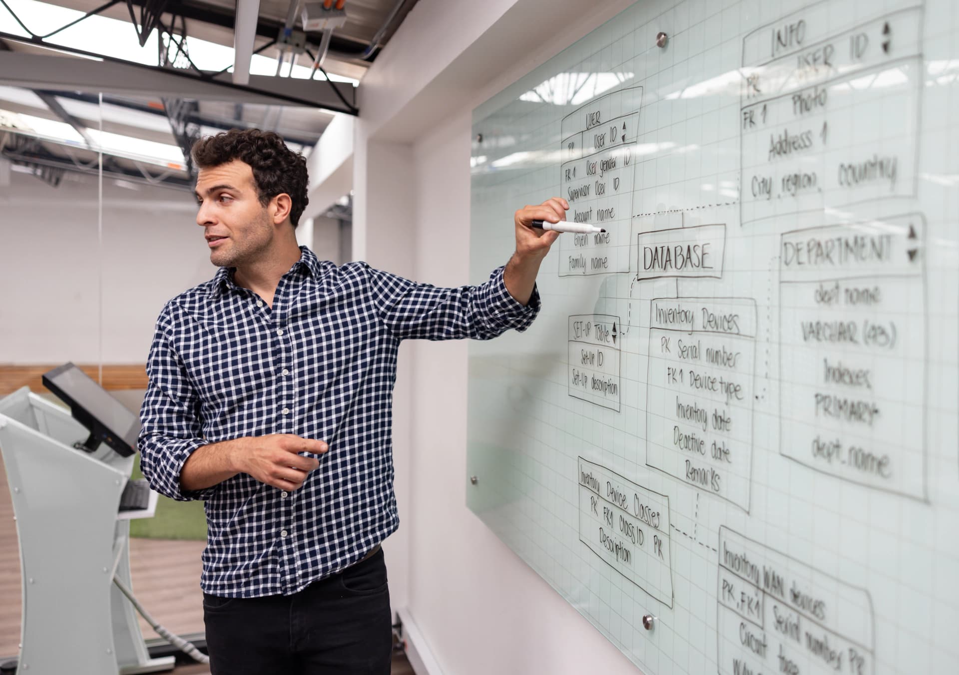 A man in a checked shirt explains a database diagram drawn on a glass board in a modern office setting.
