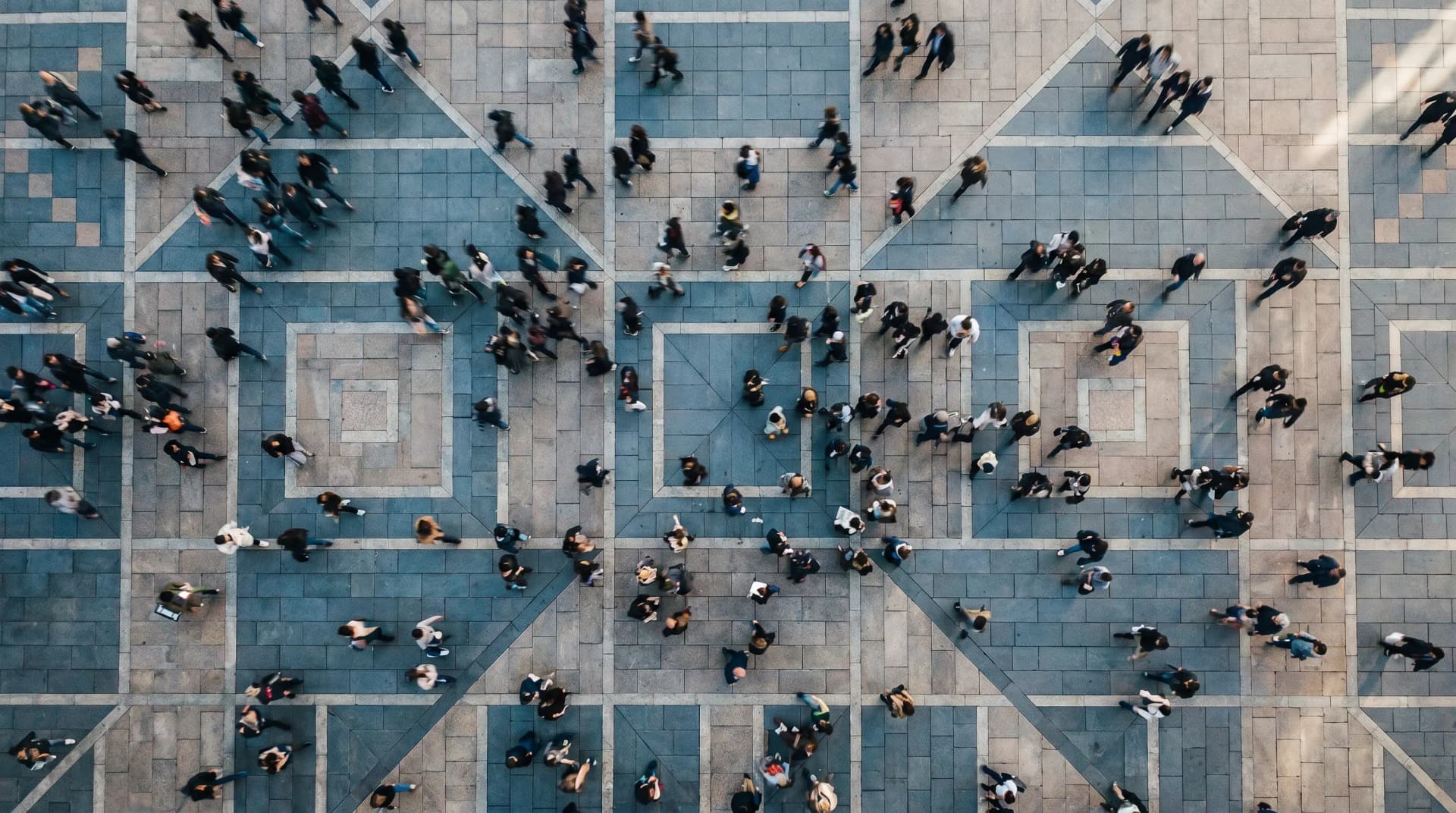 Aerial view of a crowded plaza with people walking on patterned stone tiles in various directions, creating a dynamic, bustling scene.