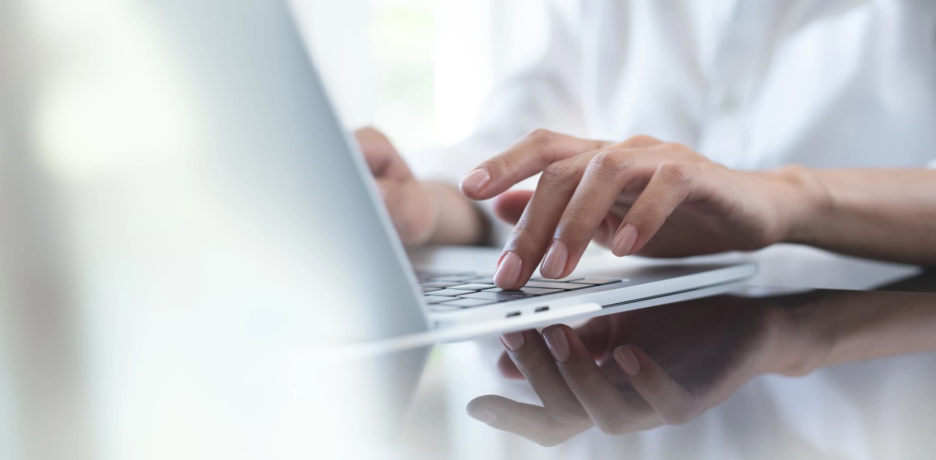 Hands typing on a laptop keyboard, with a reflection on a glossy surface, in a brightly lit setting.