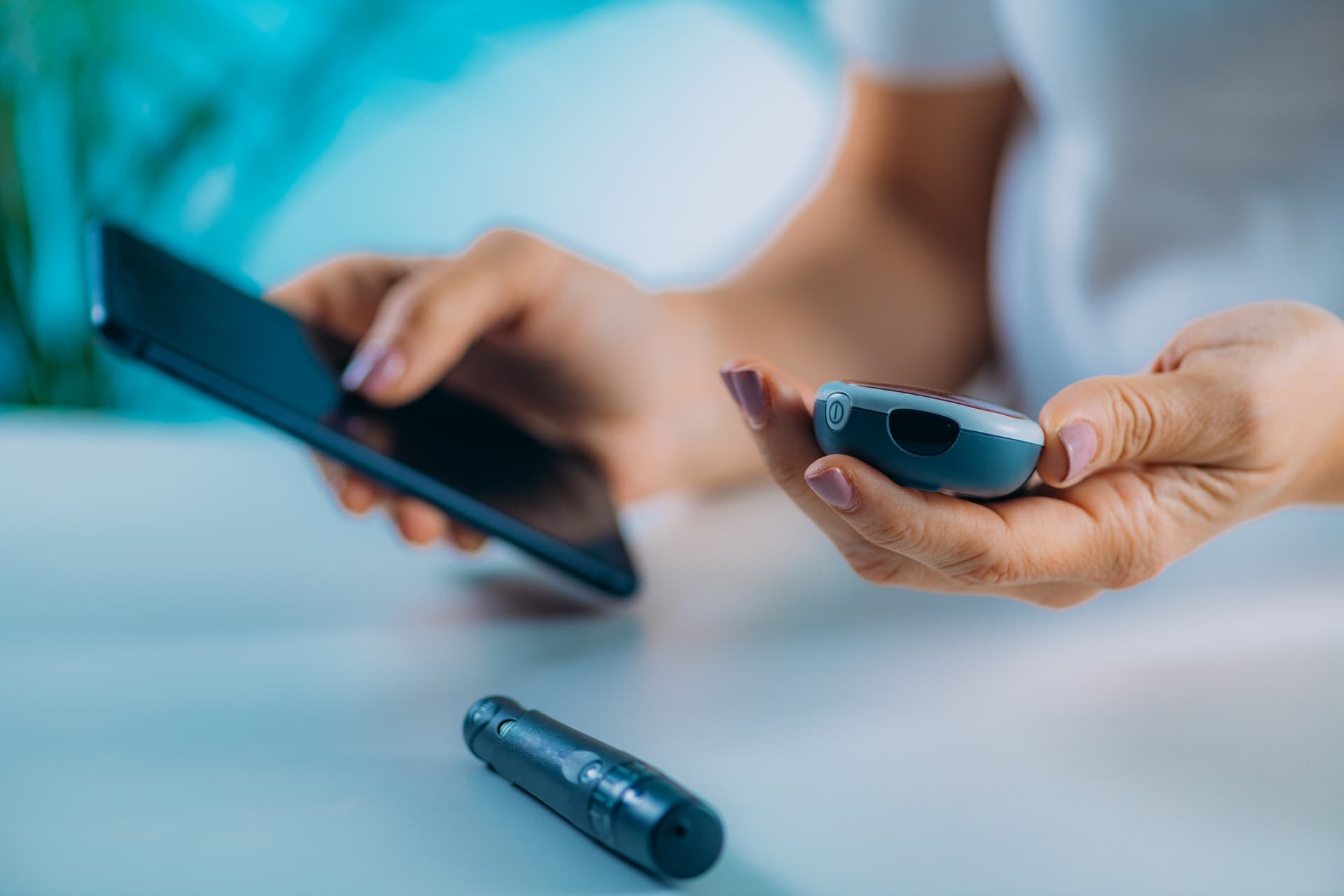 Person using a blood glucose meter and smartphone at a table, with a lancet device in the foreground.