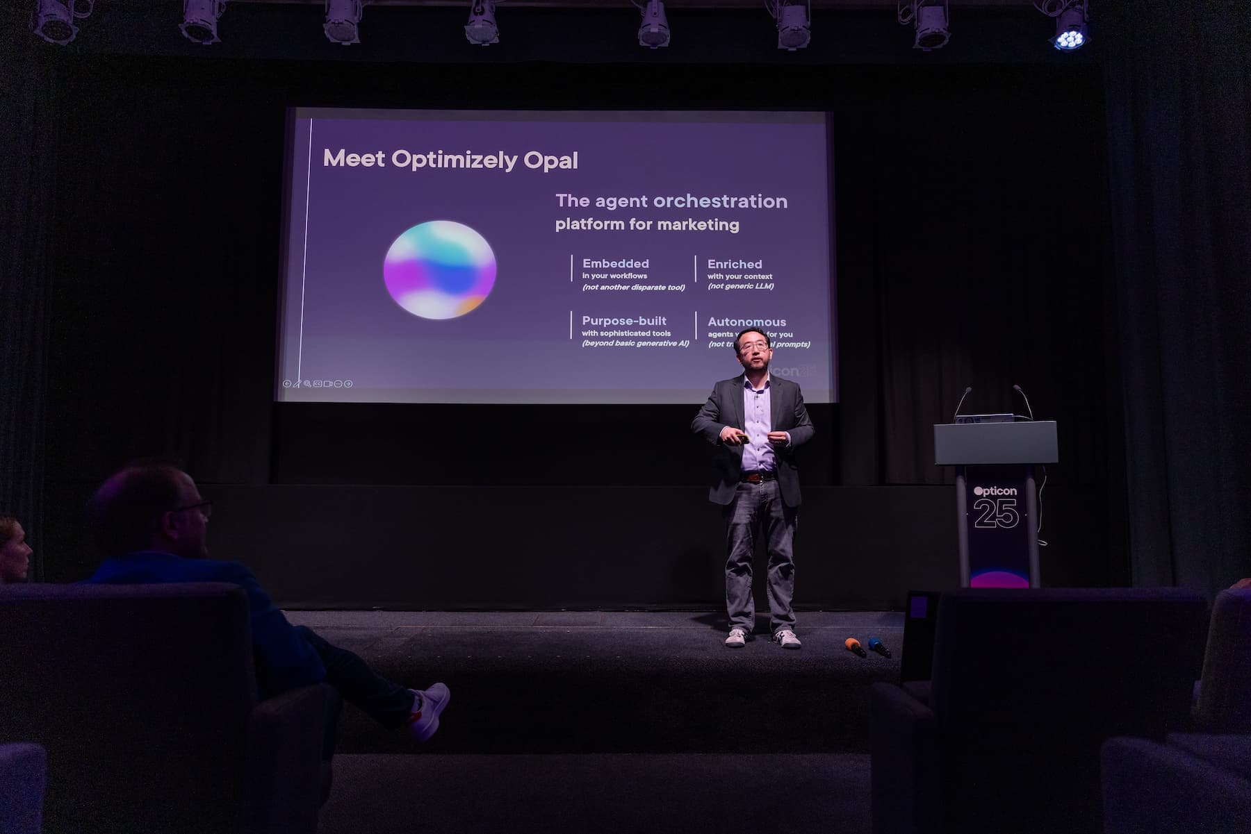 A speaker stands on stage beside a presentation slide titled "Meet Optimizely Opal" at a conference with dim lighting.