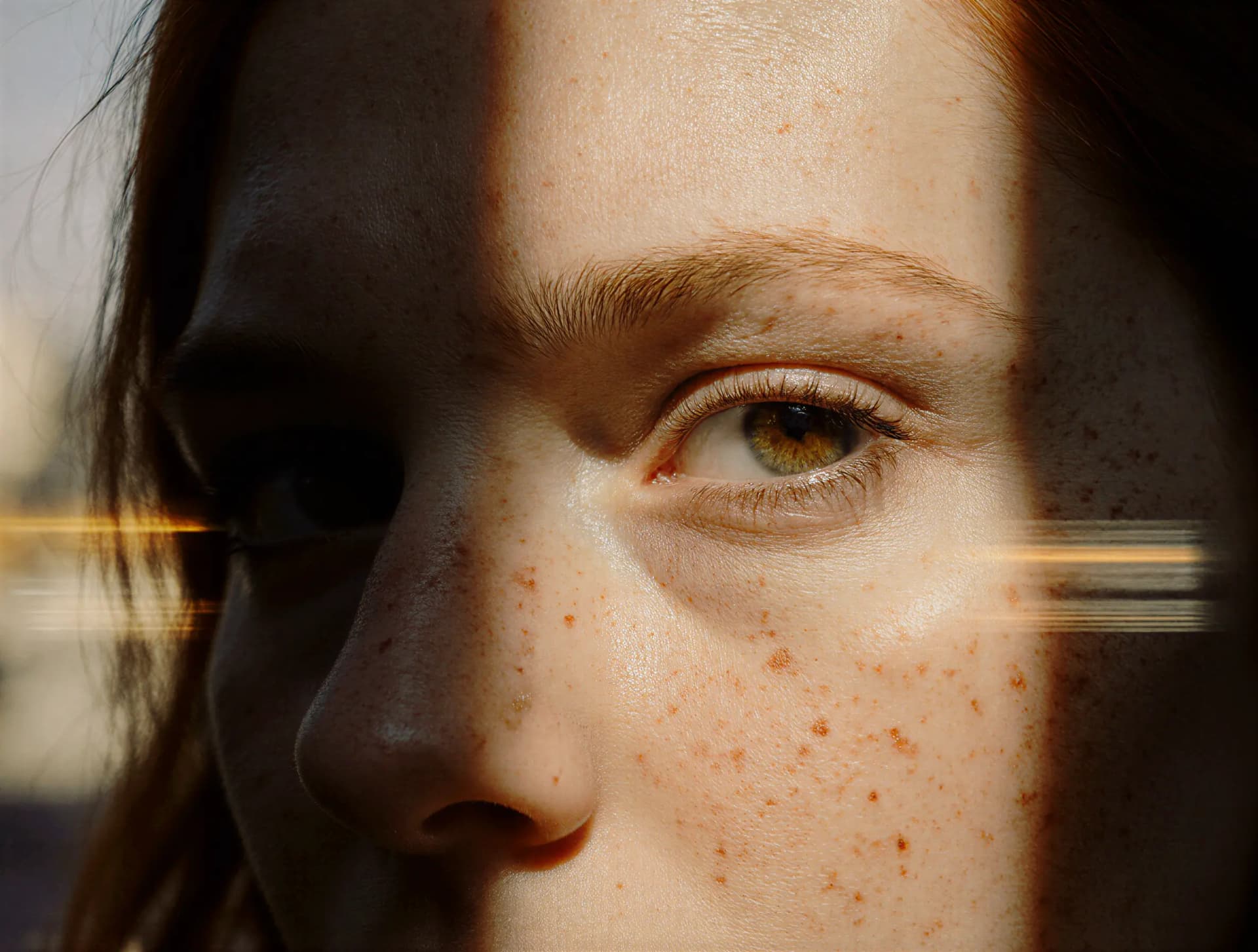 Close-up of a person's face with freckles, partially lit by sunlight, highlighting their eye and creating a shadow across their face.
