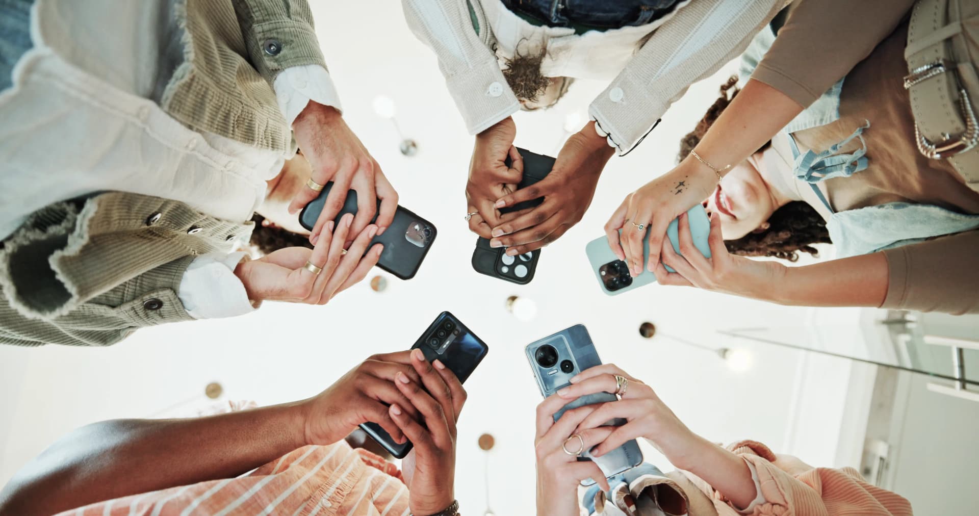 A diverse group of people standing in a circle, holding smartphones and reaching towards the center, as viewed from below.