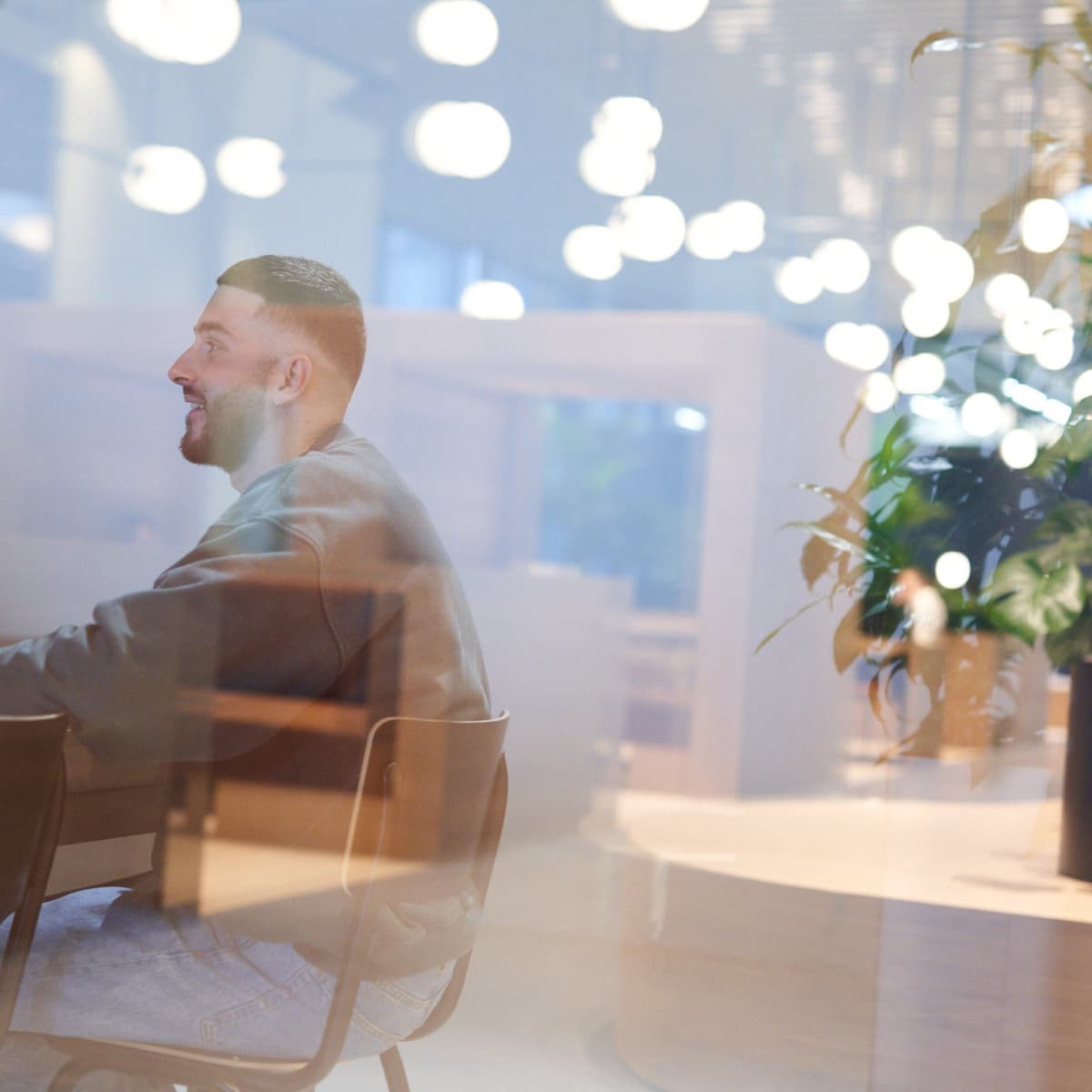 Man sitting at a table in a modern, well-lit cafe, with hanging lights and a potted plant visible through a glass window.