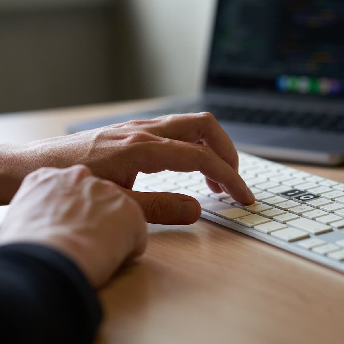 Hands typing on a white keyboard with a blurred laptop screen in the background, placed on a wooden desk.