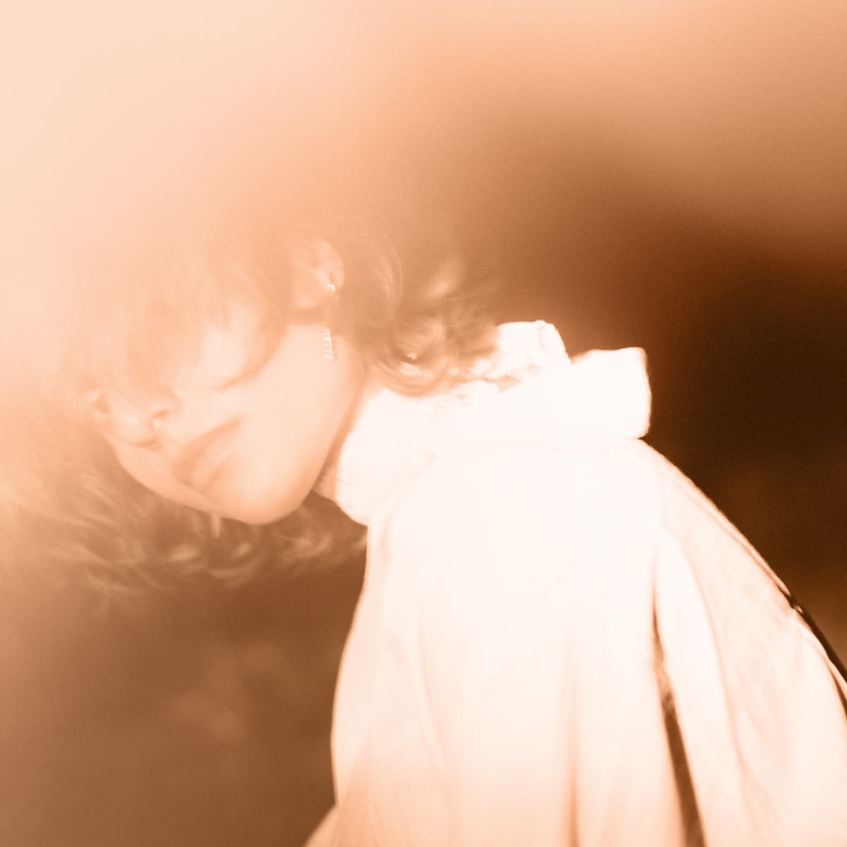 Soft-focus image of a person with a serene expression, partially obscured by a warm, hazy glow, wearing a light-colored garment.