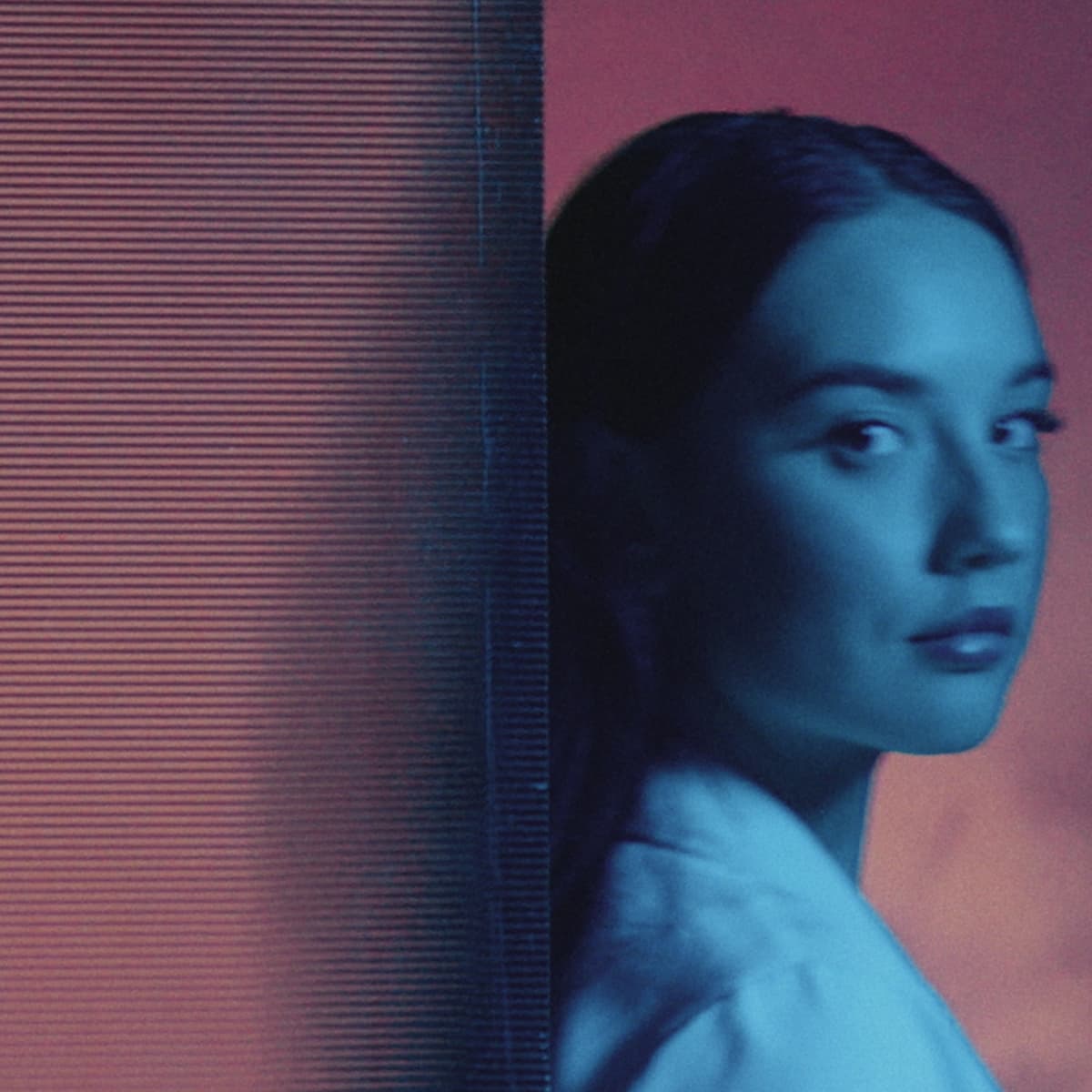 A woman with blue-toned lighting looks towards the camera, set against a pink background with vertical lines on the left side.
