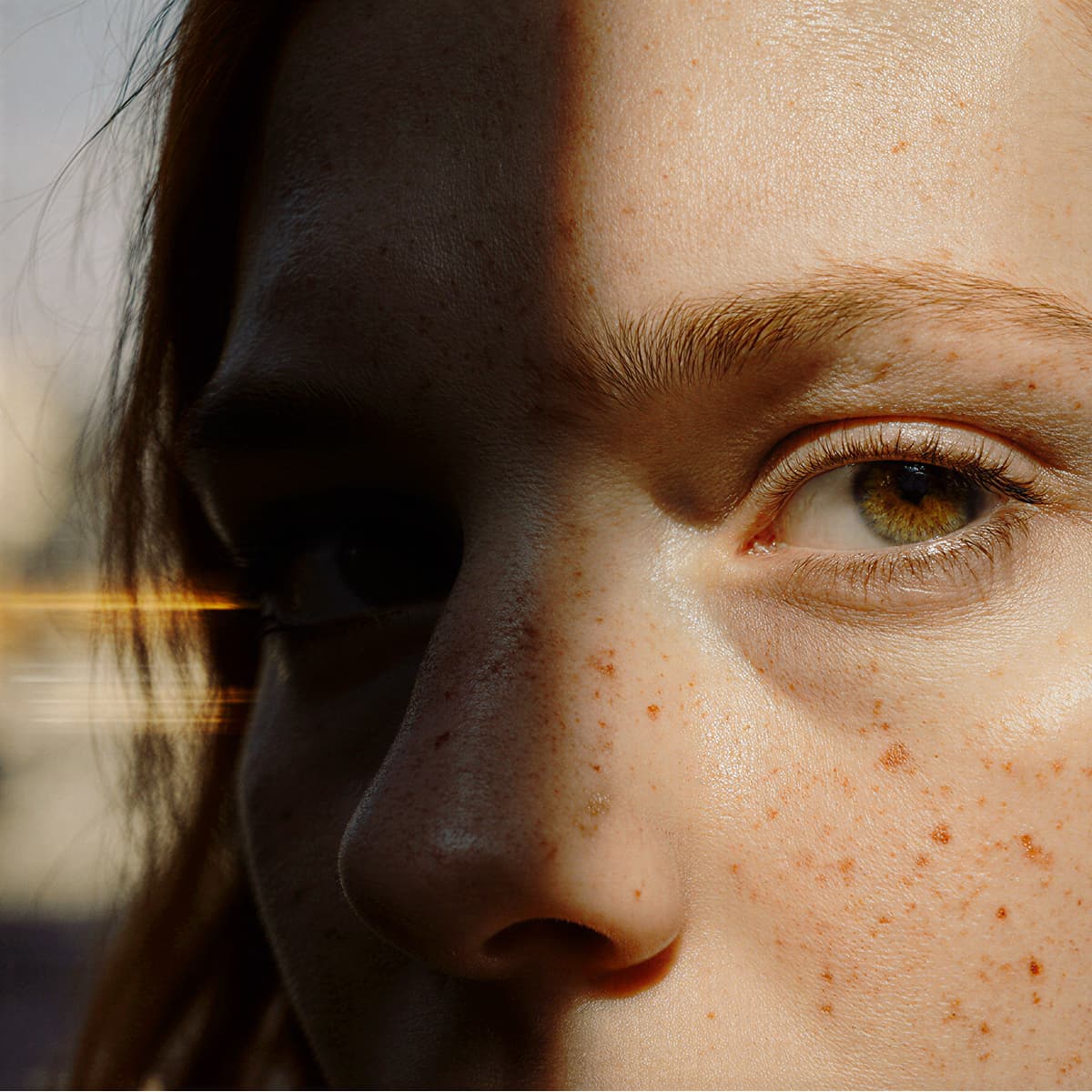 Close-up of a person's face with freckles and hazel eyes, illuminated by sunlight and shadows.