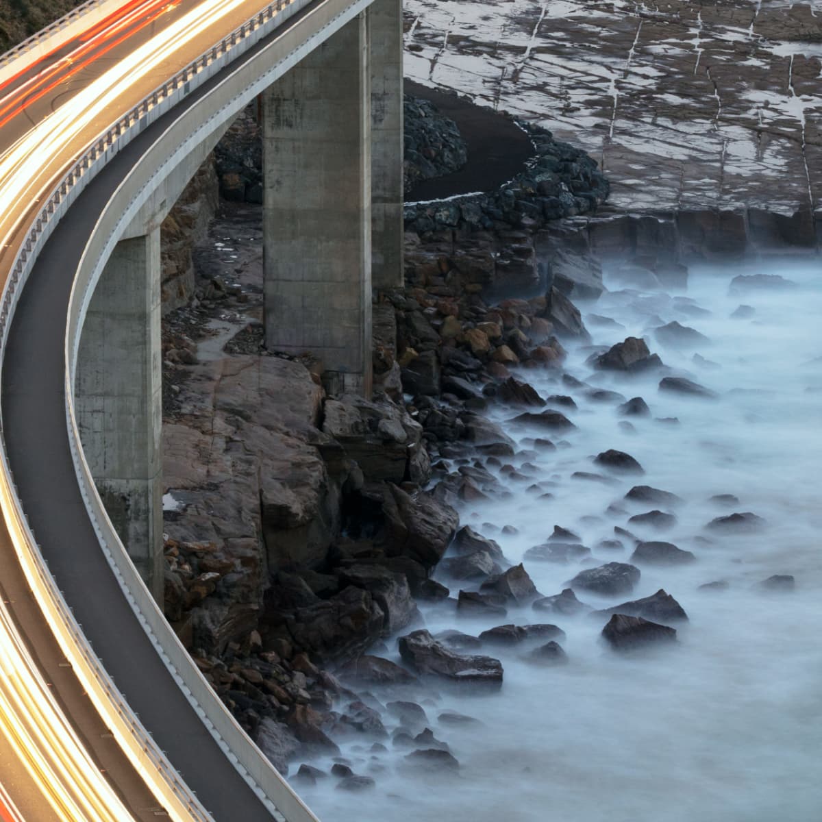 Curved coastal highway bridge with light trails, above rocky shoreline and misty waves, shot from above at dusk.