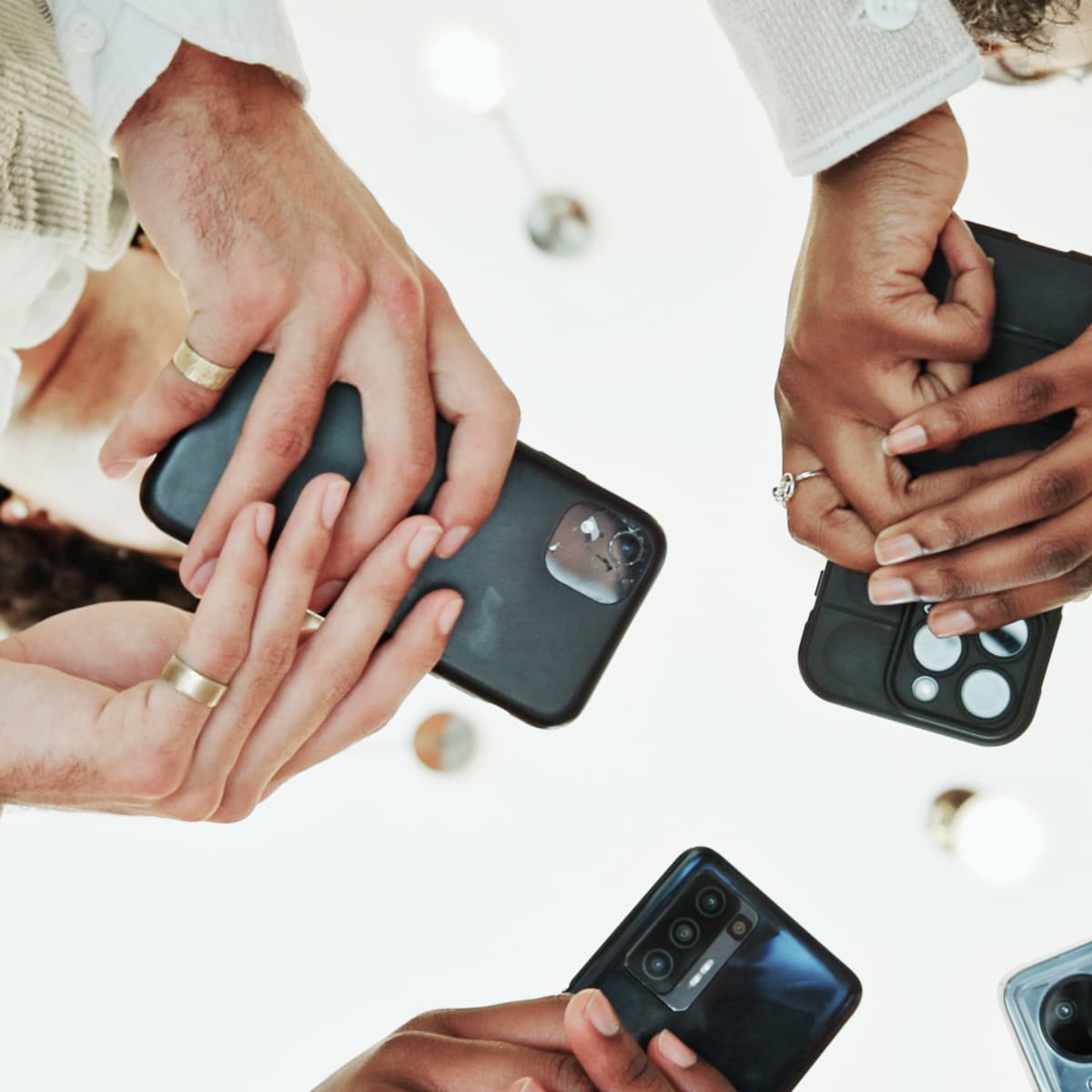 A group of hands holding various smartphones in black cases, forming a circle against a bright background.
