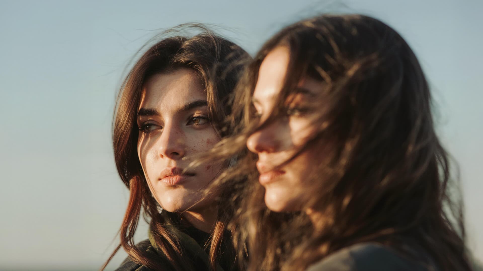 Two women with long, dark hair look into the distance, with the golden sunlight highlighting their features against a clear sky.