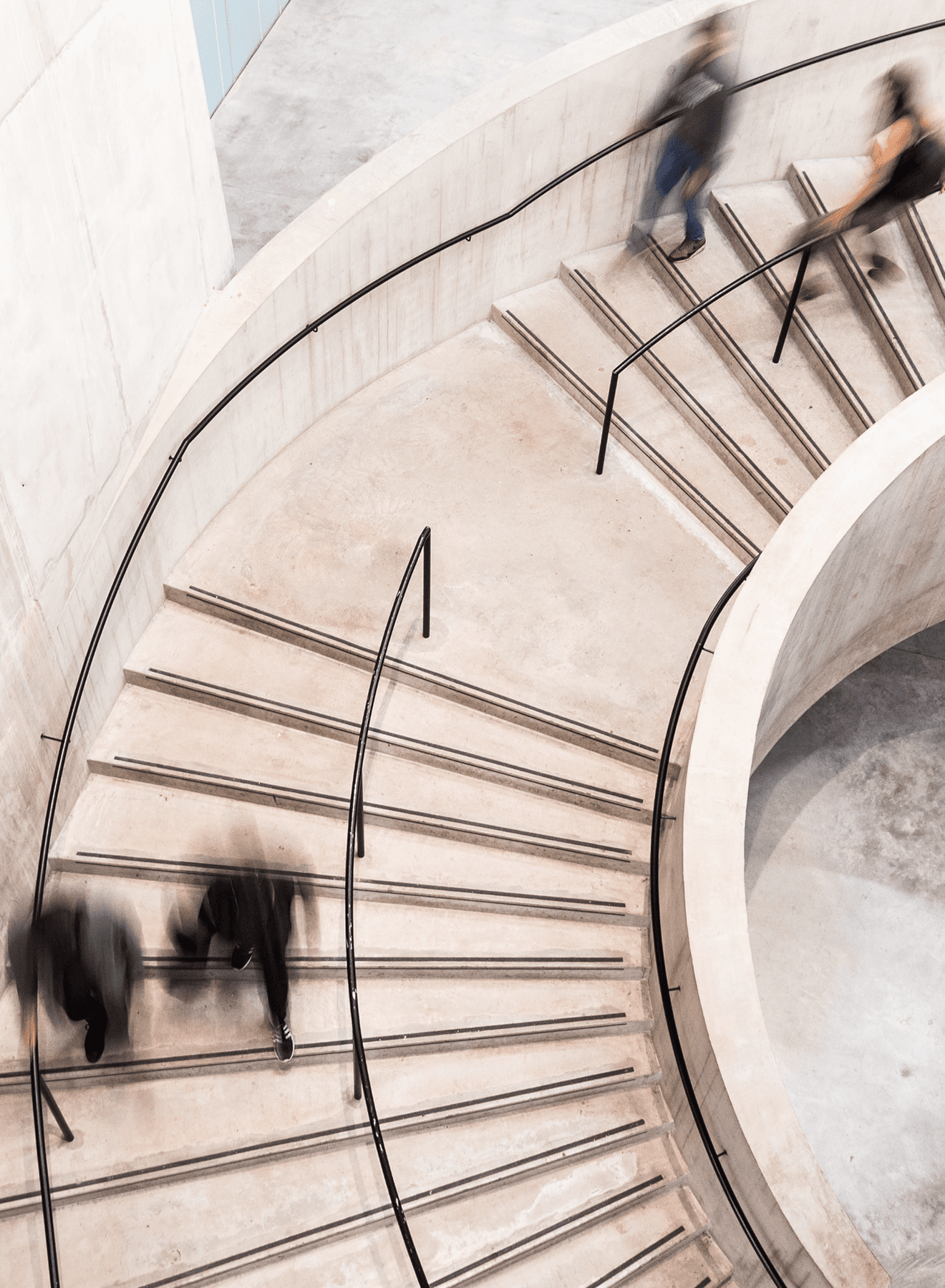 Blurred motion of people walking on a spiral staircase with concrete steps and metal railings in a modern setting.