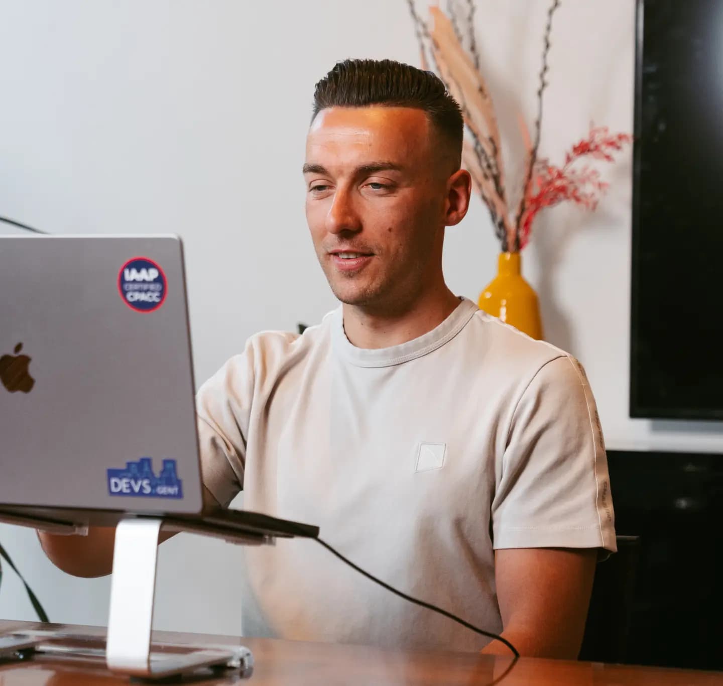 Man in a beige shirt working on a laptop with stickers, one on a stand. A yellow vase with decorative branches is in the background.