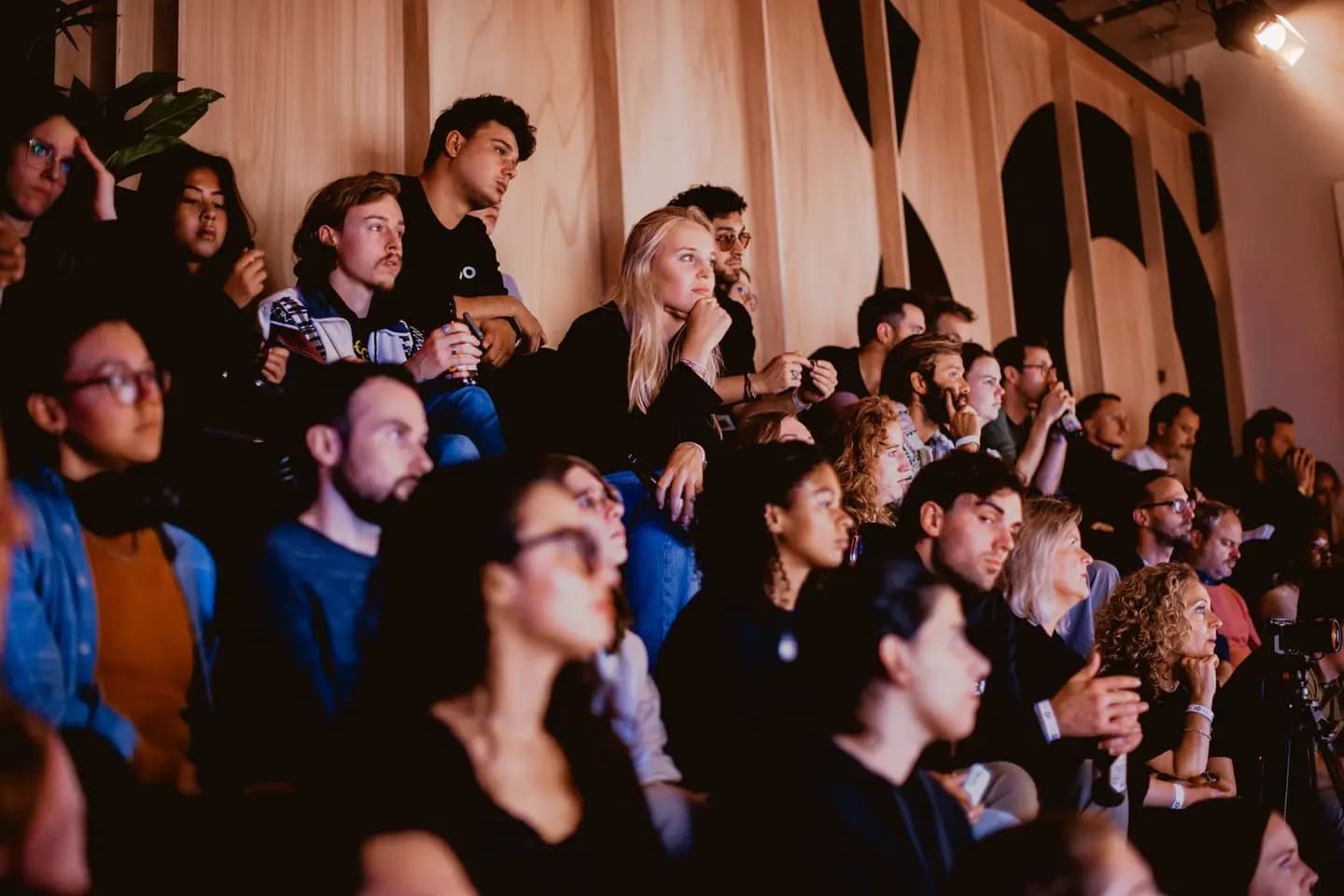 A group of people seated closely together, attentively watching an event, in a warmly lit auditorium with wooden panel walls.